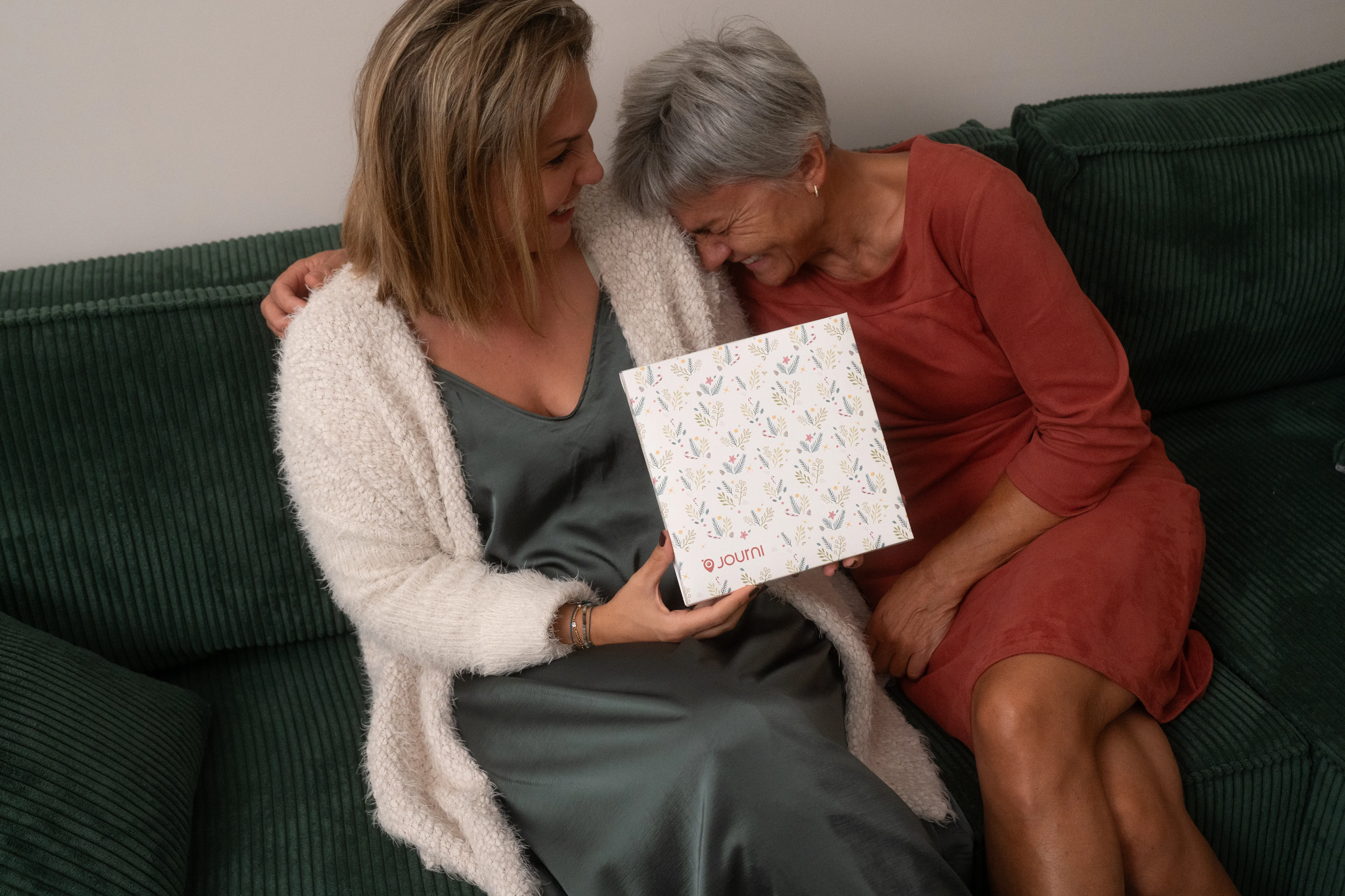 Zwei Frauen sitzen auf einem grünen Sofa, lächeln und halten gemeinsam eine gemusterte Geschenkbox in den Händen.