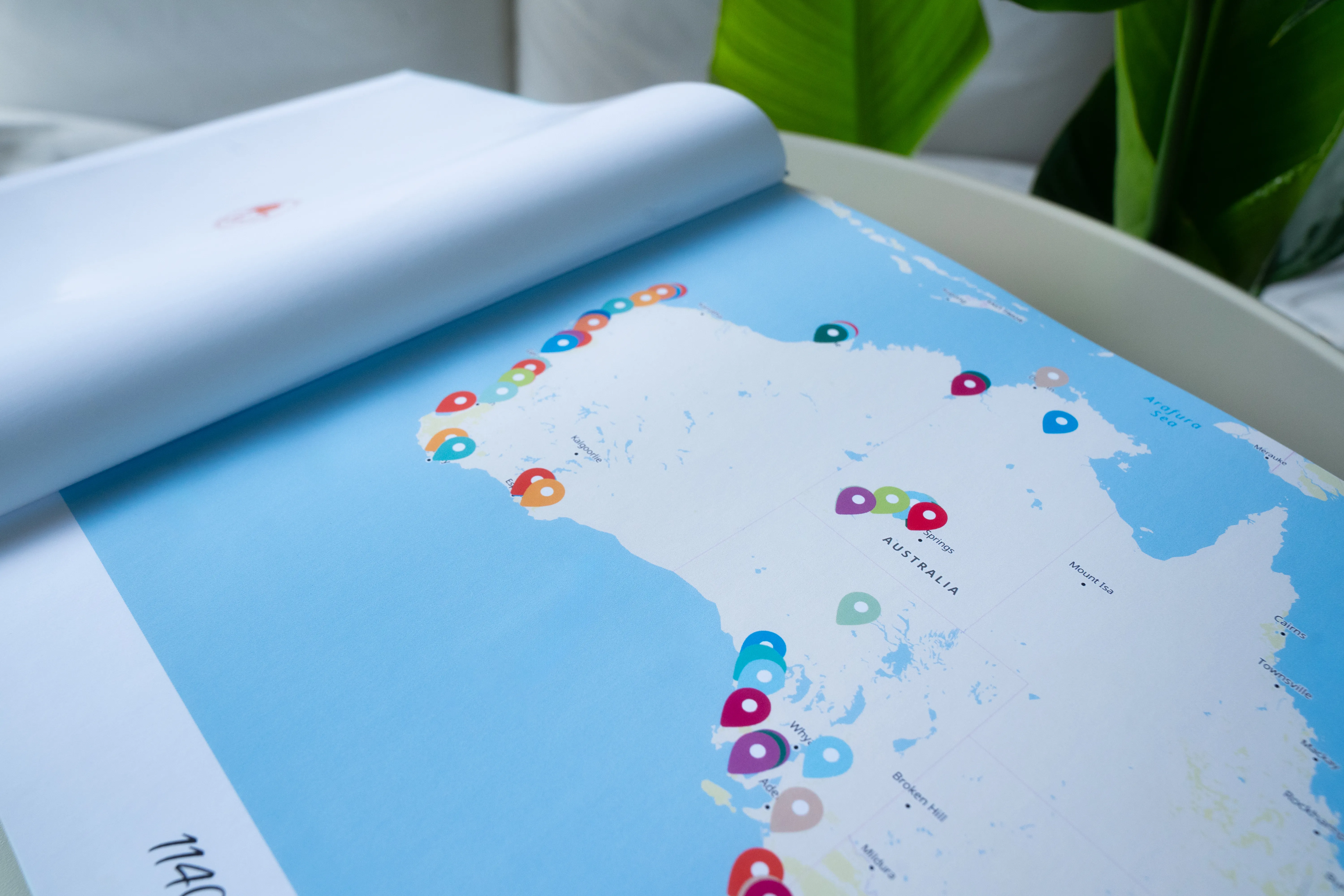 Open travel photo book showing a map of Australia with colorful location markers, placed on a light surface near green leaves.