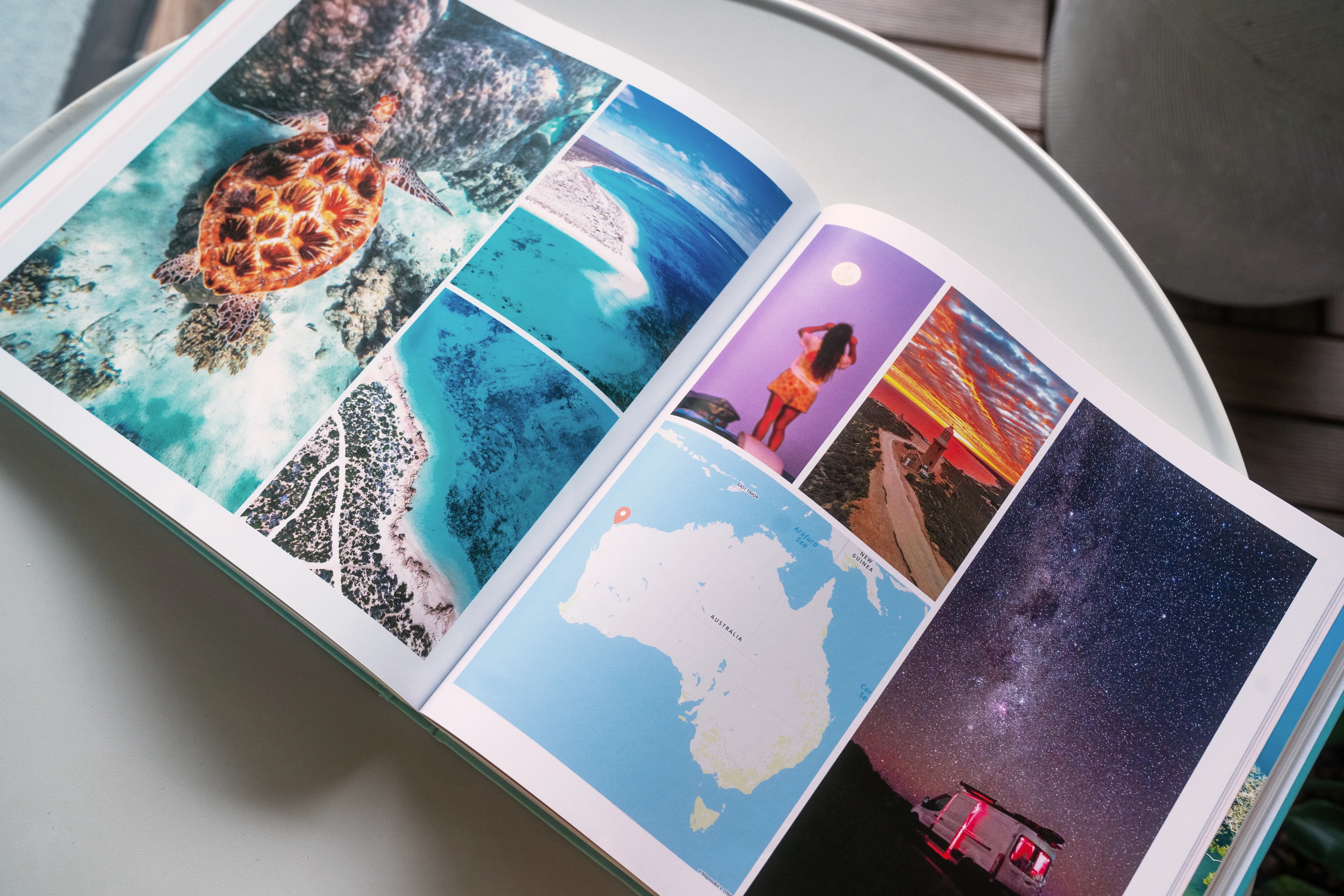 Ein aufgeschlagenes Reisefotobuch mit farbenfrohen Urlaubsfotos – darunter eine Schildkröte, Strände, ein Nachthimmel und eine Karte von Australien – liegt auf einem weißen Tisch.