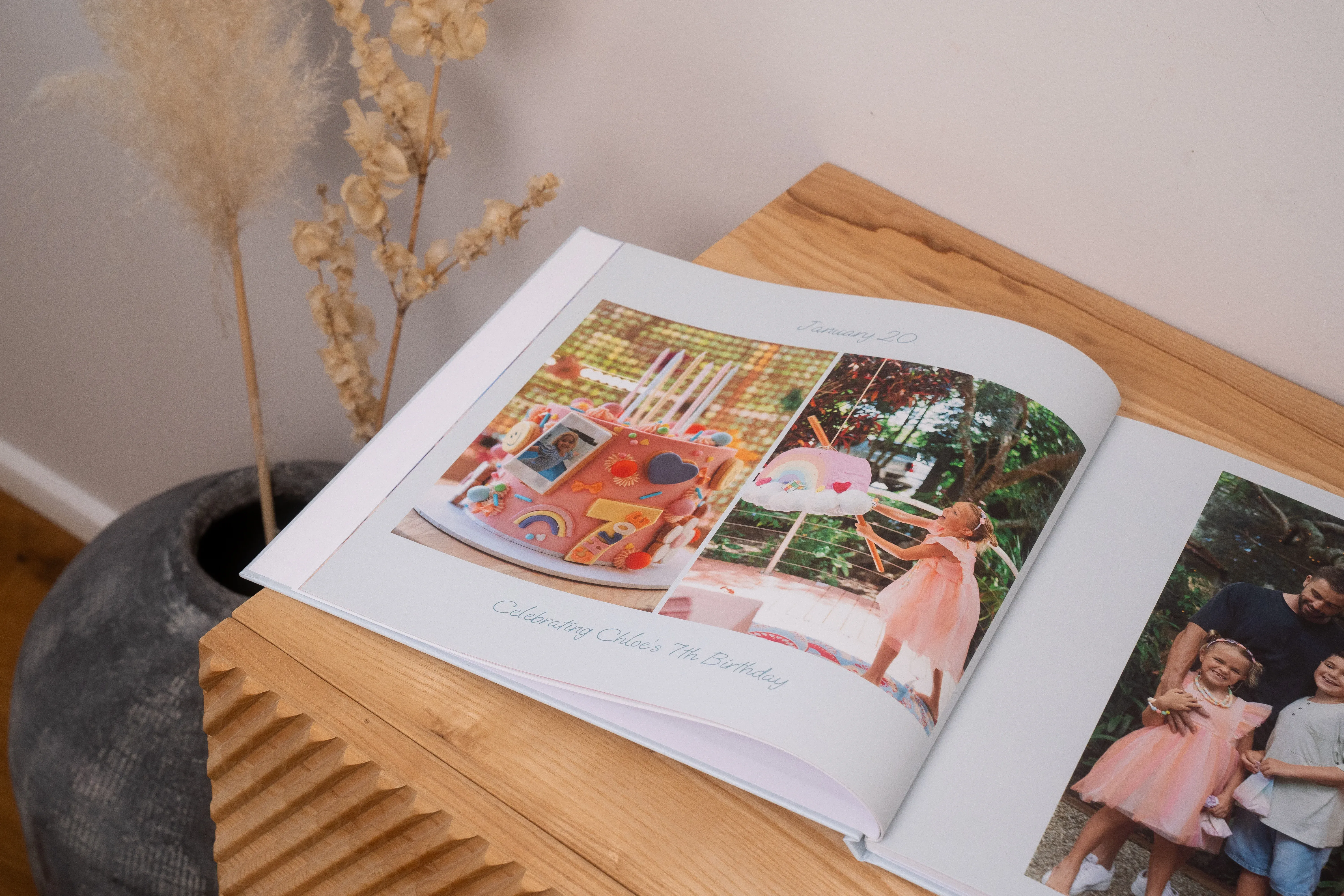 Ein offenes Geburtstags-Fotobuch auf einem Holzregal neben einer Pflanze. Es sind Fotos eines Kindergeburtstages zu sehen.