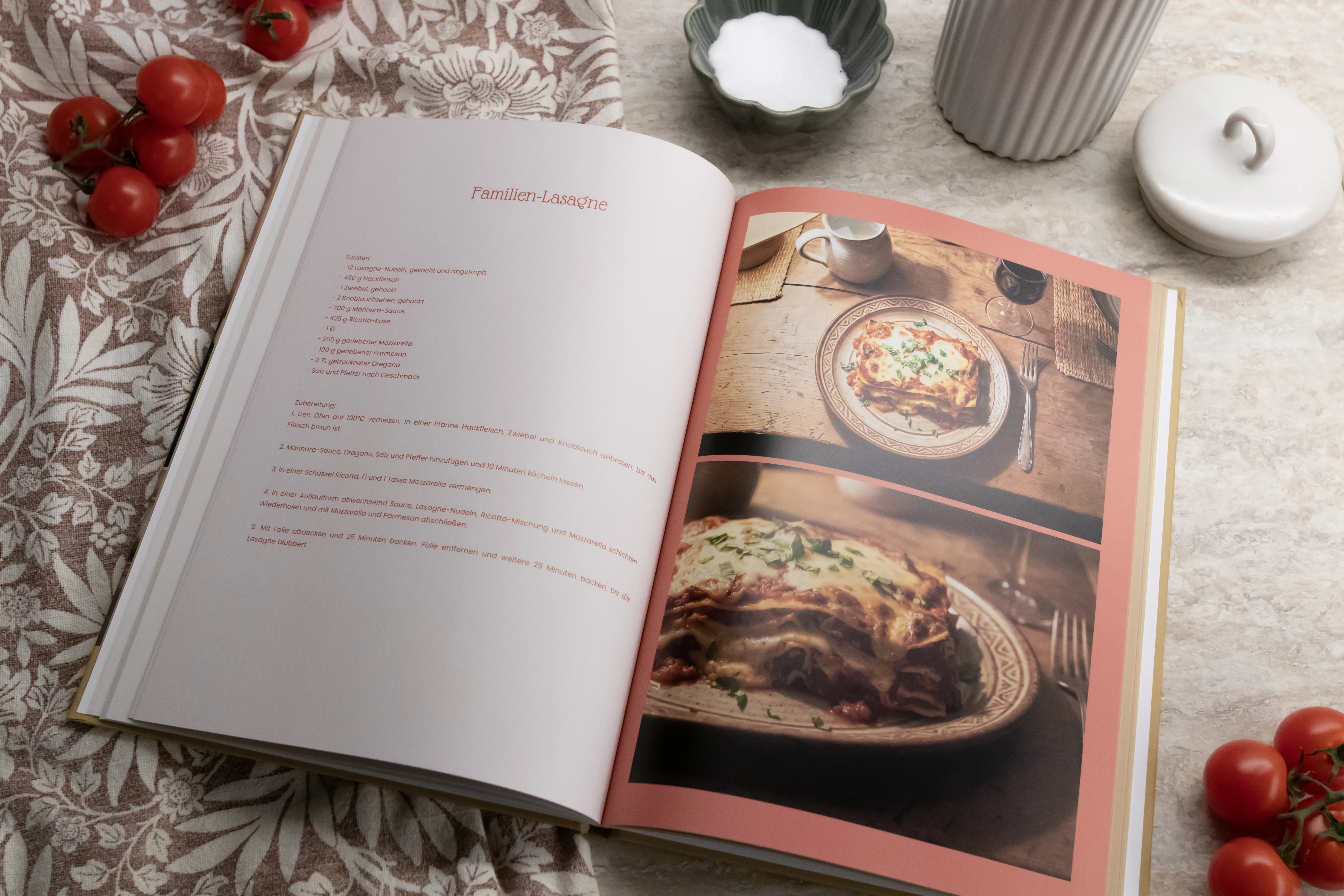 Open cookbook photo book on a tablecloth, featuring a lasagna recipe with accompanying photos, surrounded by fresh tomatoes and kitchenware.