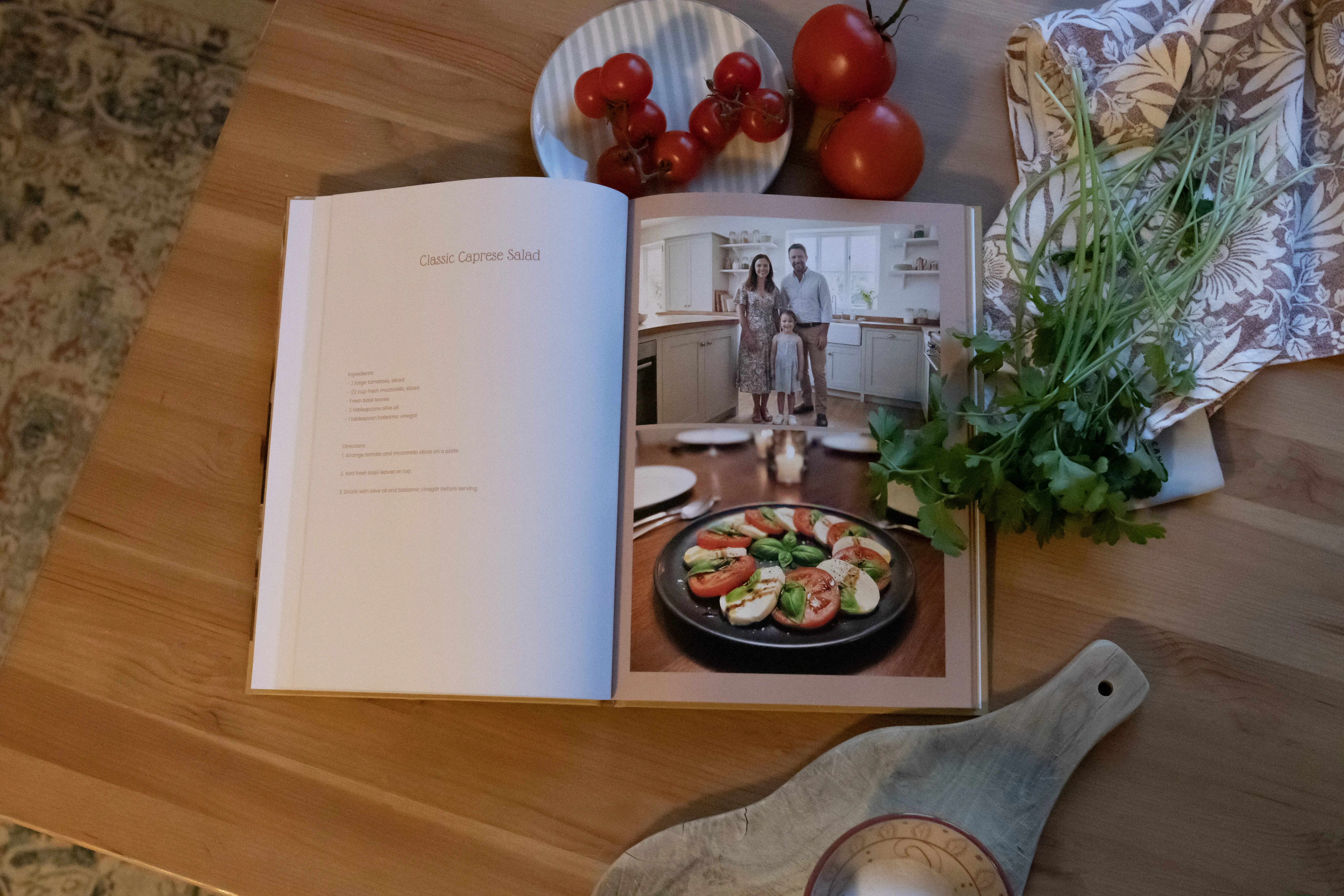 Open cookbook on a wooden table with a caprese salad photo, cherry tomatoes, herbs, and patterned fabric nearby.