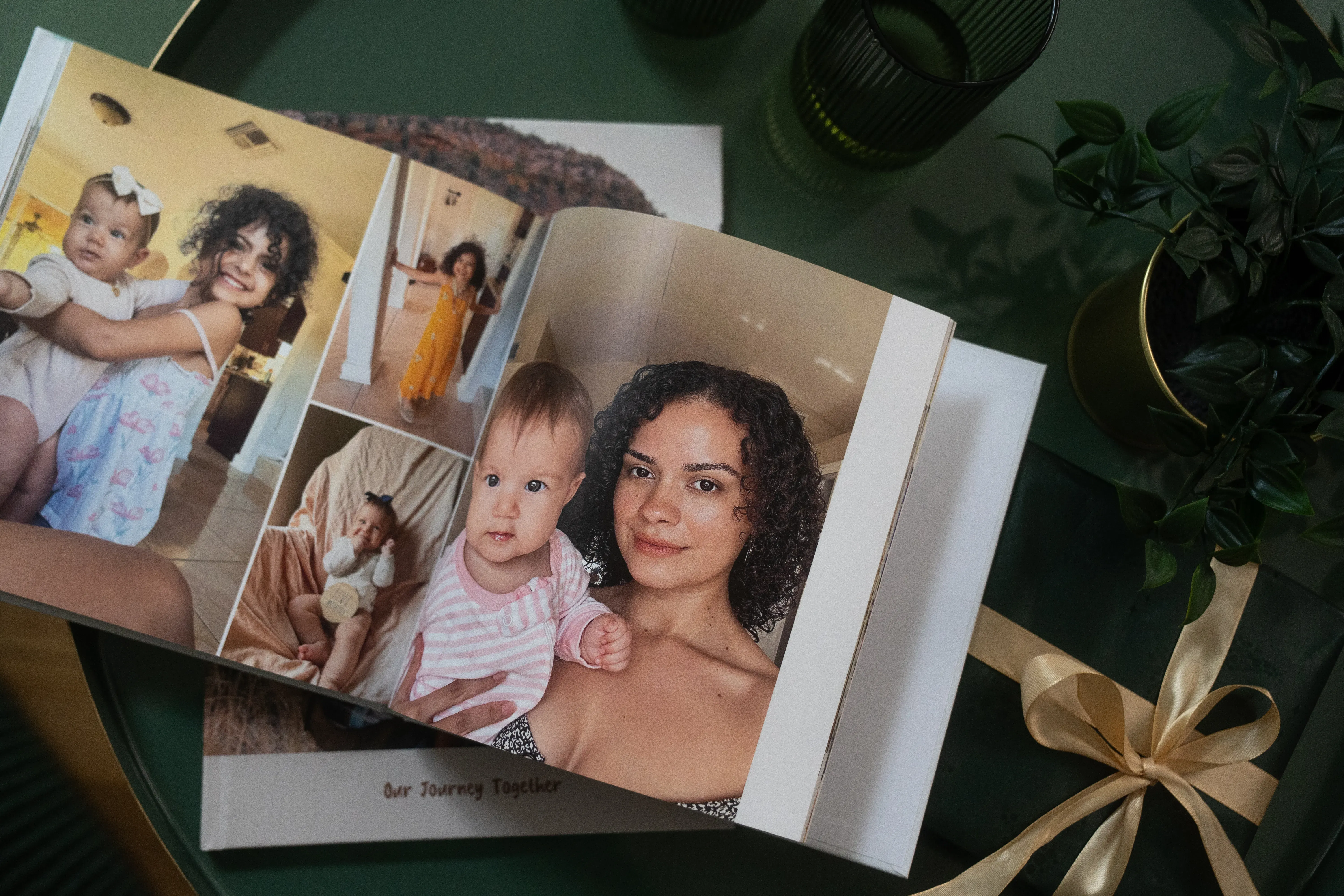 Open baby photo album on a table with pictures of a woman and baby; nearby are plants and a ribbon-tied gift box.