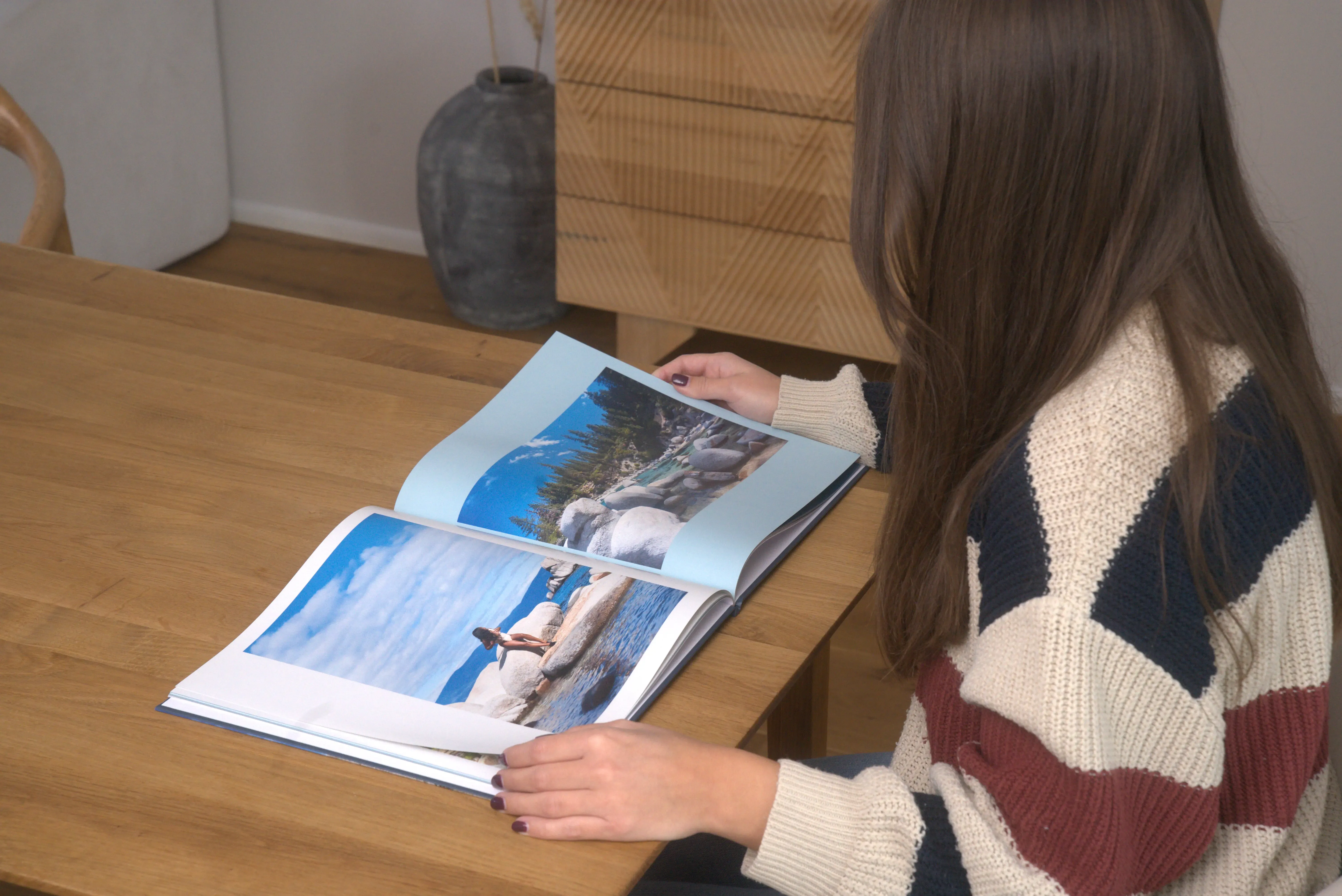 A woman with long hair in a striped sweater looks through a photo book on a wooden table, featuring beach and nature images.