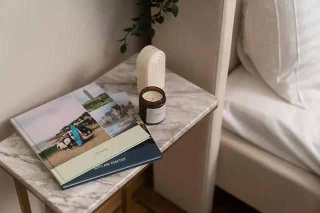 An elegant collection of hardcover travel photo books displayed as a premium home decor piece on a chic marble bedside table, styled beautifully alongside a glowing scented candle next to crisp white bed linens.