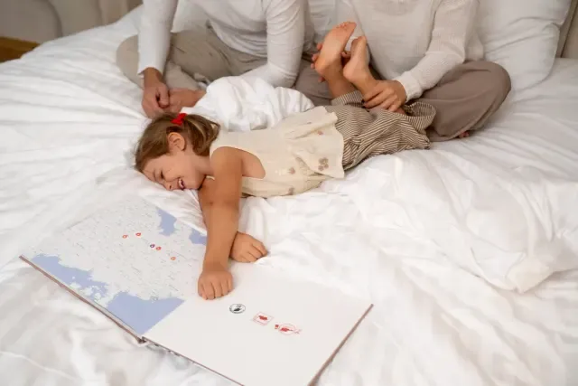 Child lying on a bed with a layflat photo book open, smiling and playing while two adults sit nearby.