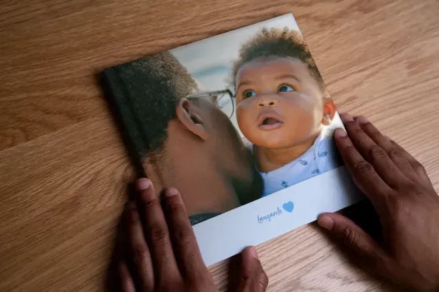Hands holding a layflat photo book with a cover showing a person kissing a baby's cheek, placed on a wooden surface.
