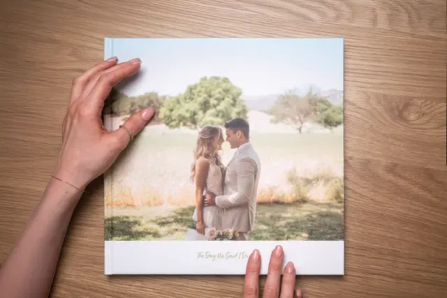 Person holding a wedding photo album with a cover featuring a couple embracing in a serene outdoor setting, with trees in the background.