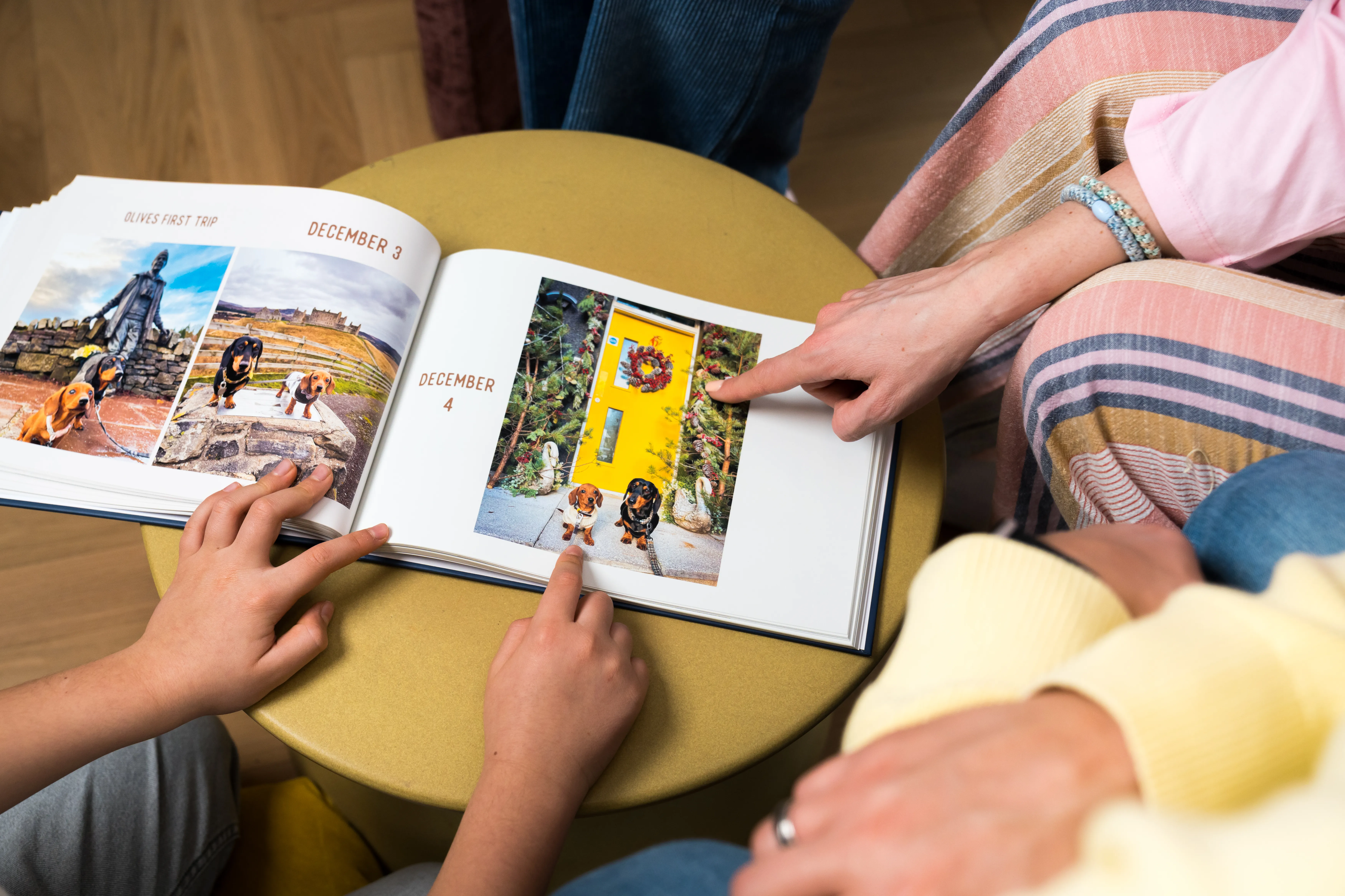 People pointing at photos in an open book on a table, featuring colorful outdoor scenes with animals on December pages.
