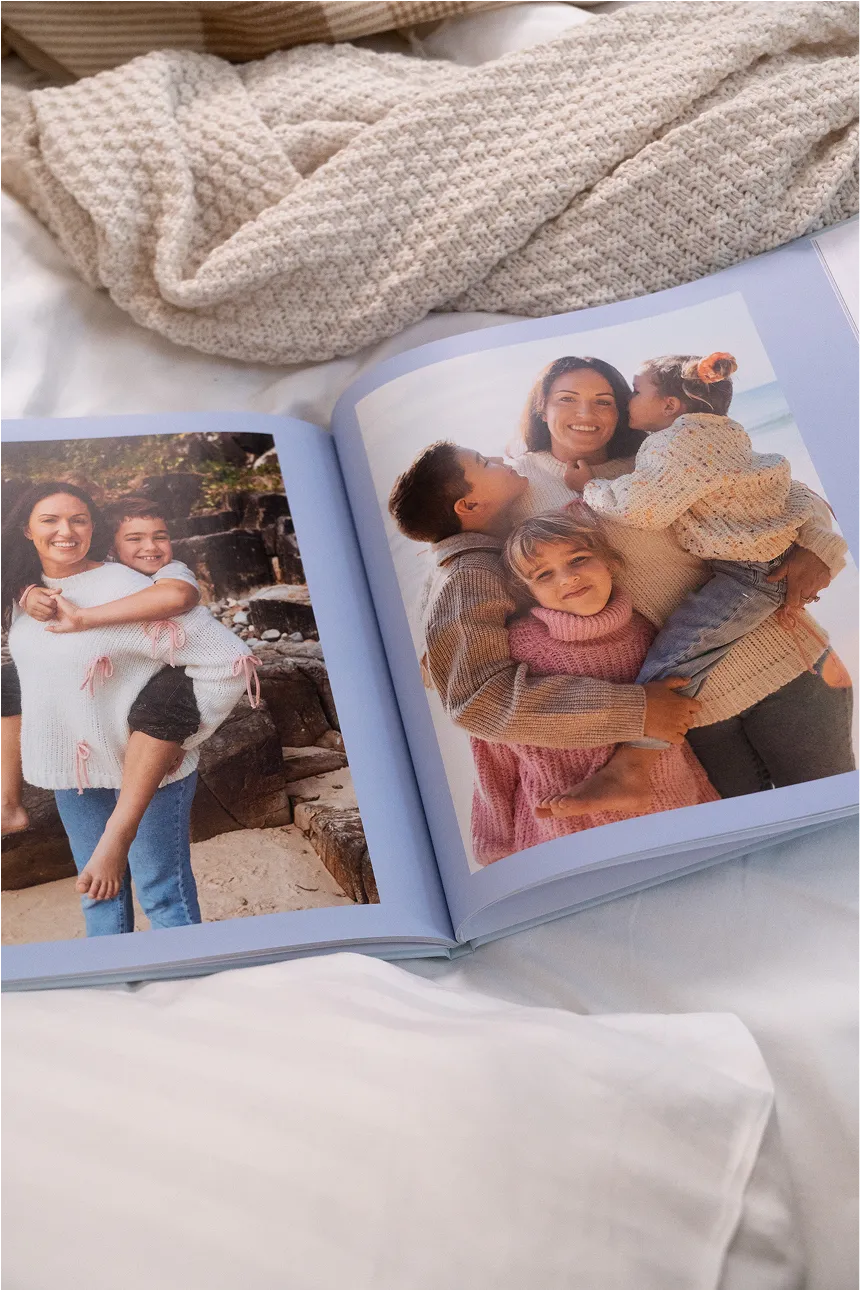 Open photo album showing a woman with three children outdoors, smiling and hugging, placed on a cozy bed with a knitted blanket.