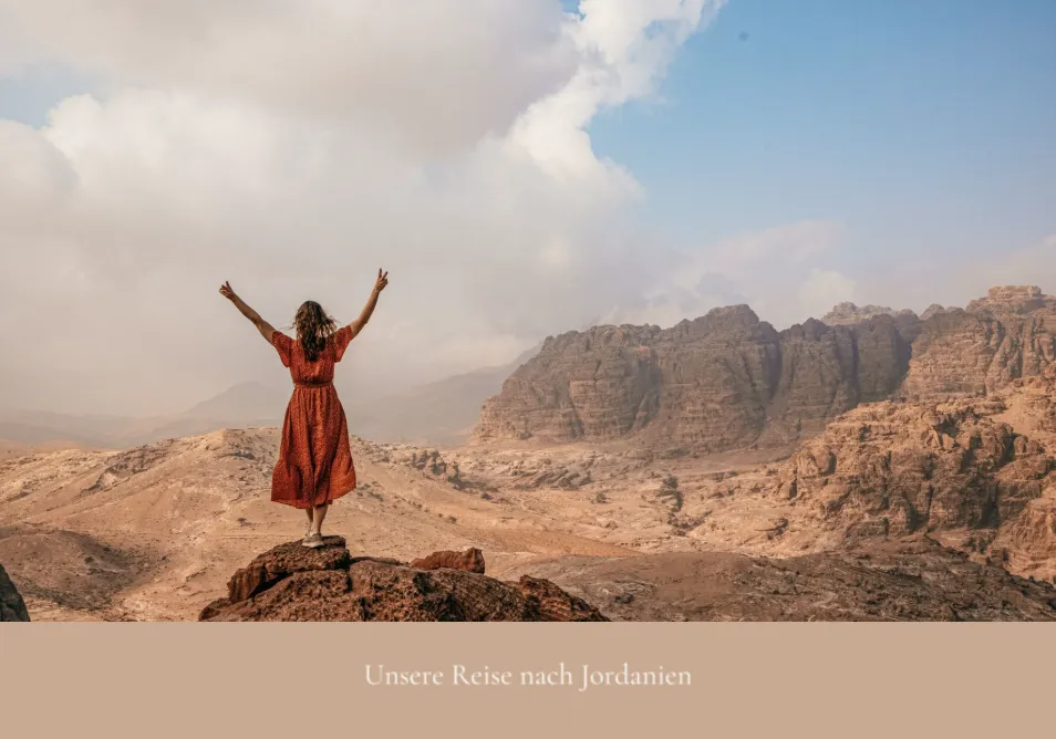 Eine Person in einem roten Kleid steht mit erhobenen Armen auf einer felsigen Klippe und schaut auf eine riesige Wüstenlandschaft unter einem bewölkten Himmel.