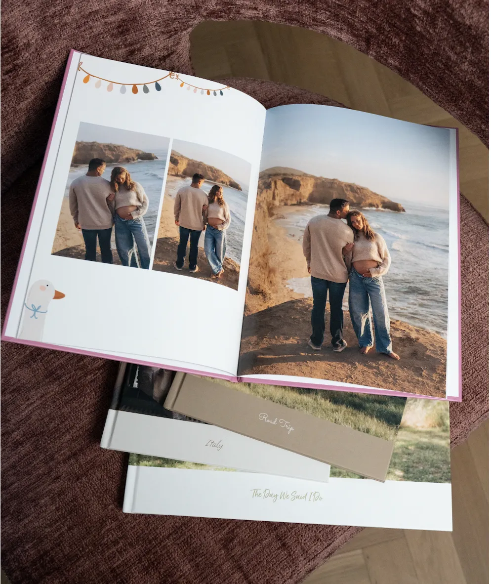 Ein offenes Fotobuch auf einer strukturierten roten Fläche, auf dem Bilder eines Paares an einer Klippe am Meer zu sehen sind; darunter liegen weitere Alben.