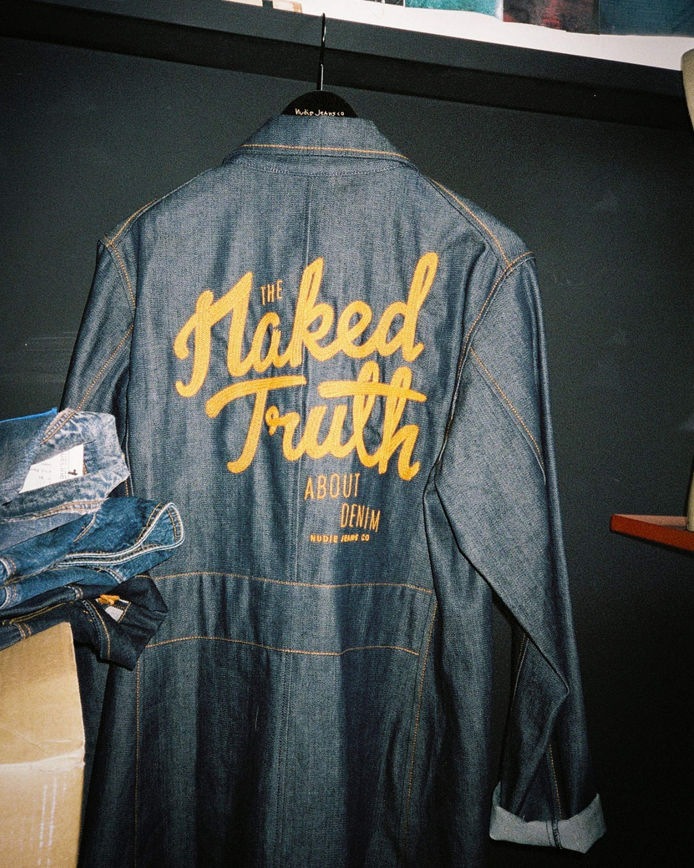Denim jacket hanging, featuring bold yellow text: "The Naked Truth About Denim" on the back, with a stack of jeans nearby.