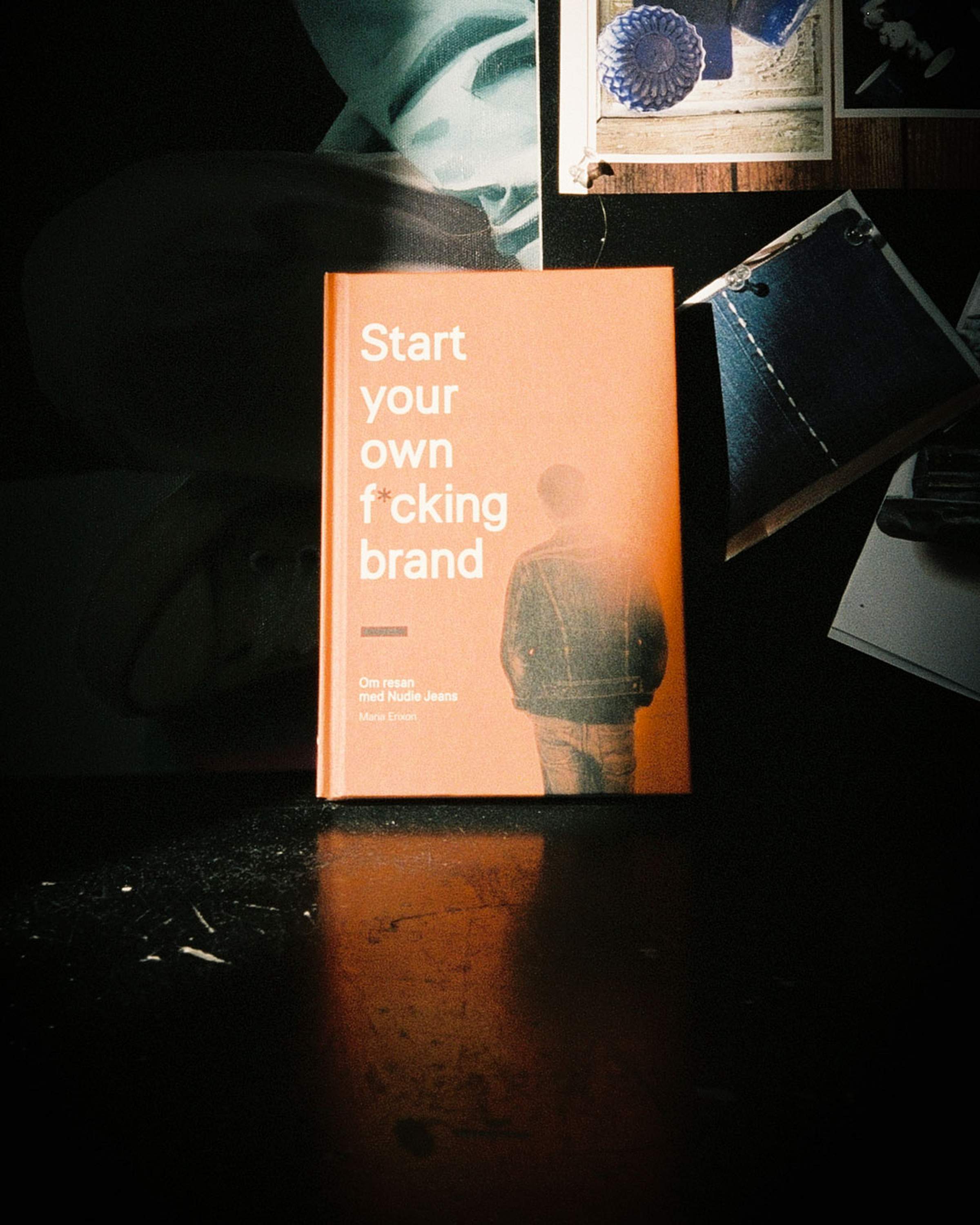 Book titled "Start your own f*cking brand" on a dark surface, surrounded by photos and fabric swatches.