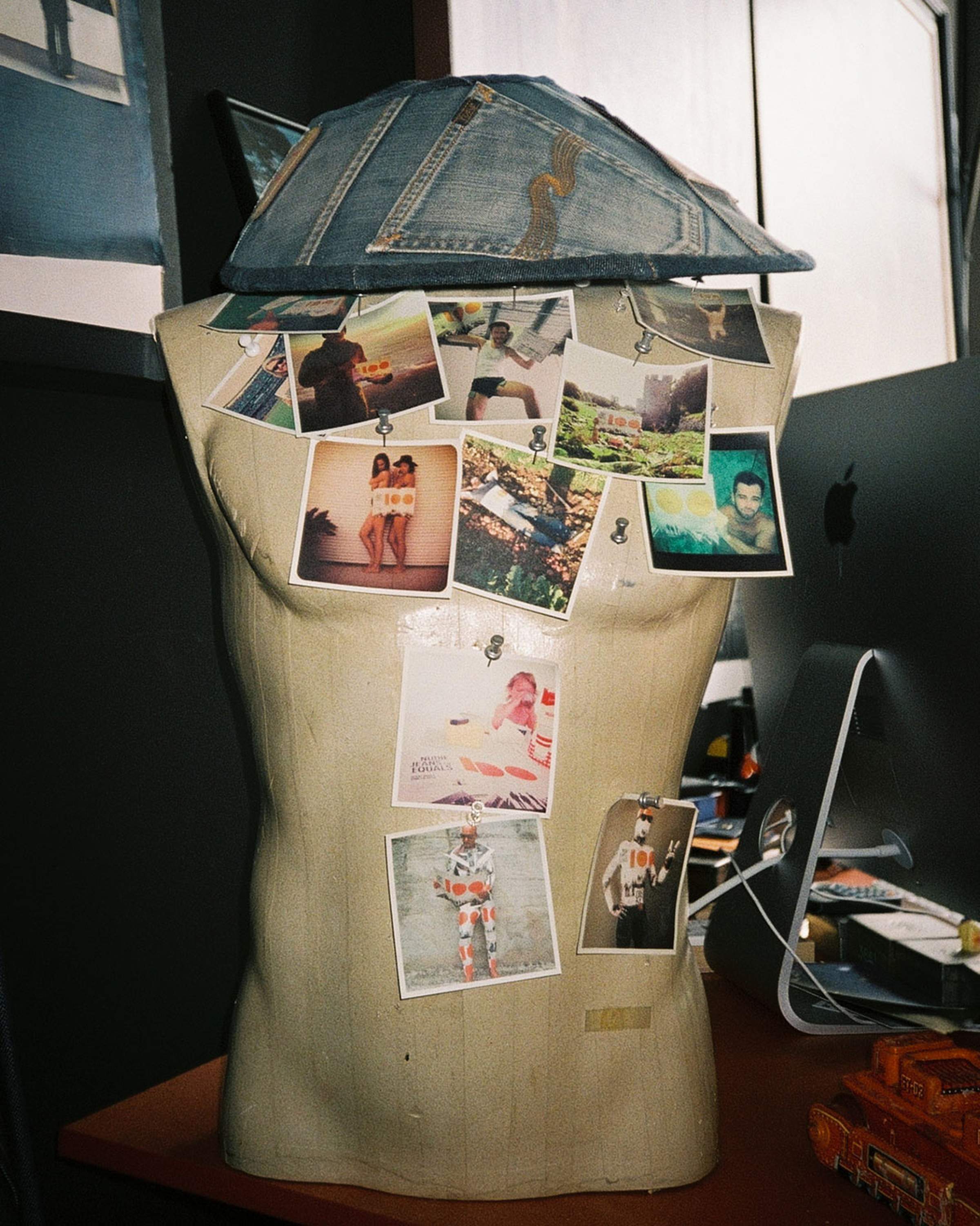 Mannequin torso with denim hat, adorned with various pinned photos, stands beside a computer on a desk.