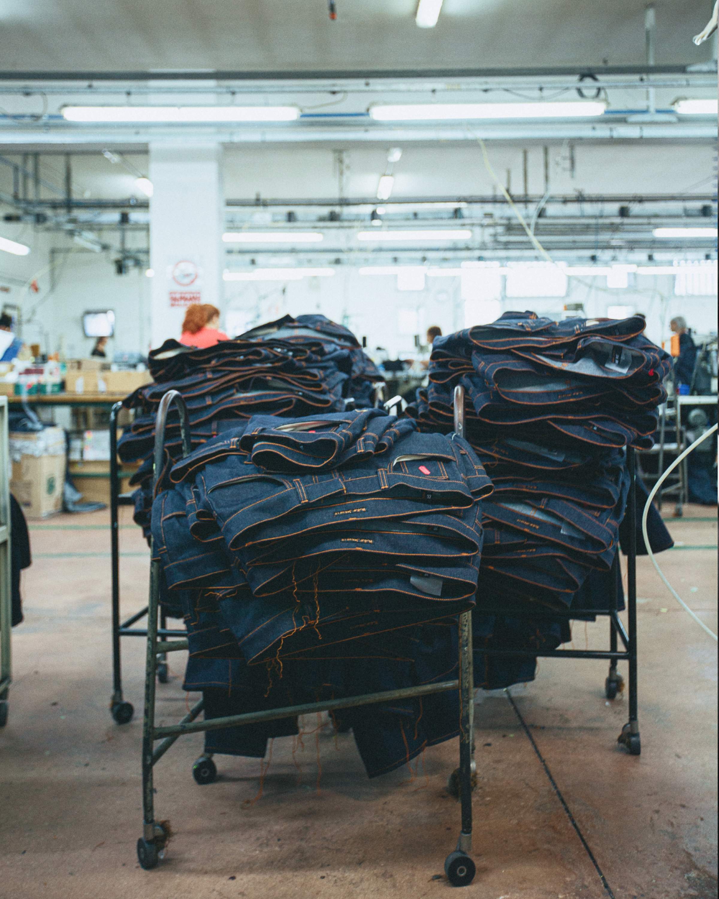 Stacks of dry, raw Nudie Jeans on metal carts in a factory setting, with workers in the background.