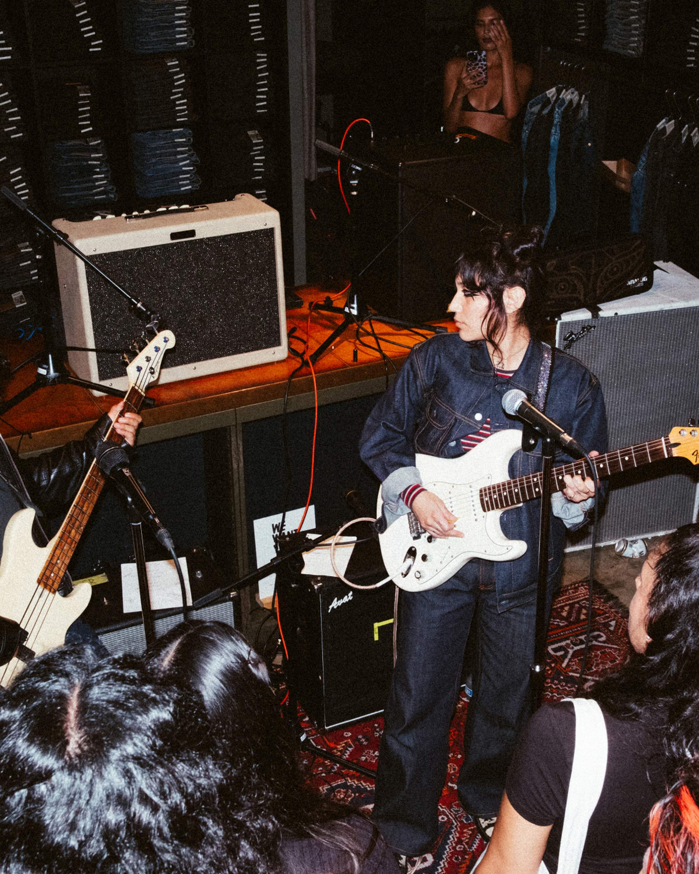 Musicians perform with guitars and amplifiers in a small venue, surrounded by attendees. A person in the background captures the moment on a phone.