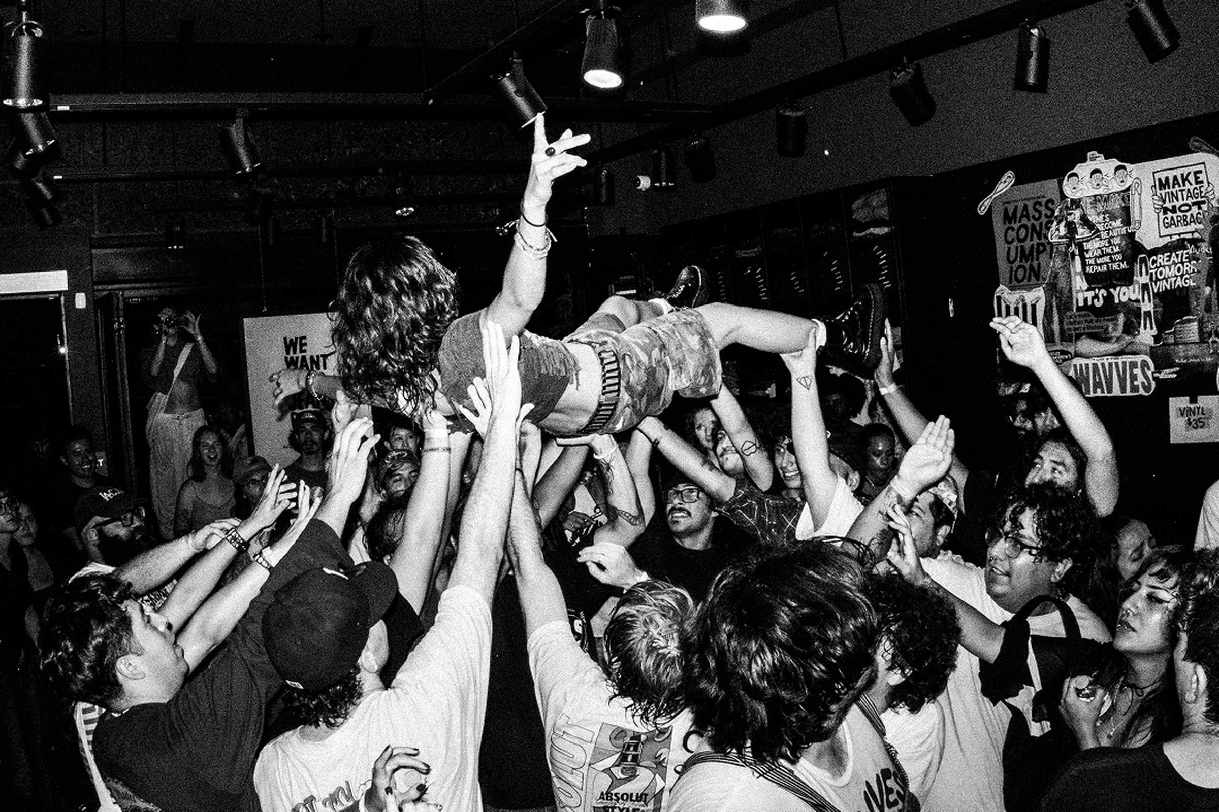 A crowd of excited concertgoers supports a person crowd-surfing in a lively, packed indoor venue, with posters in the background.