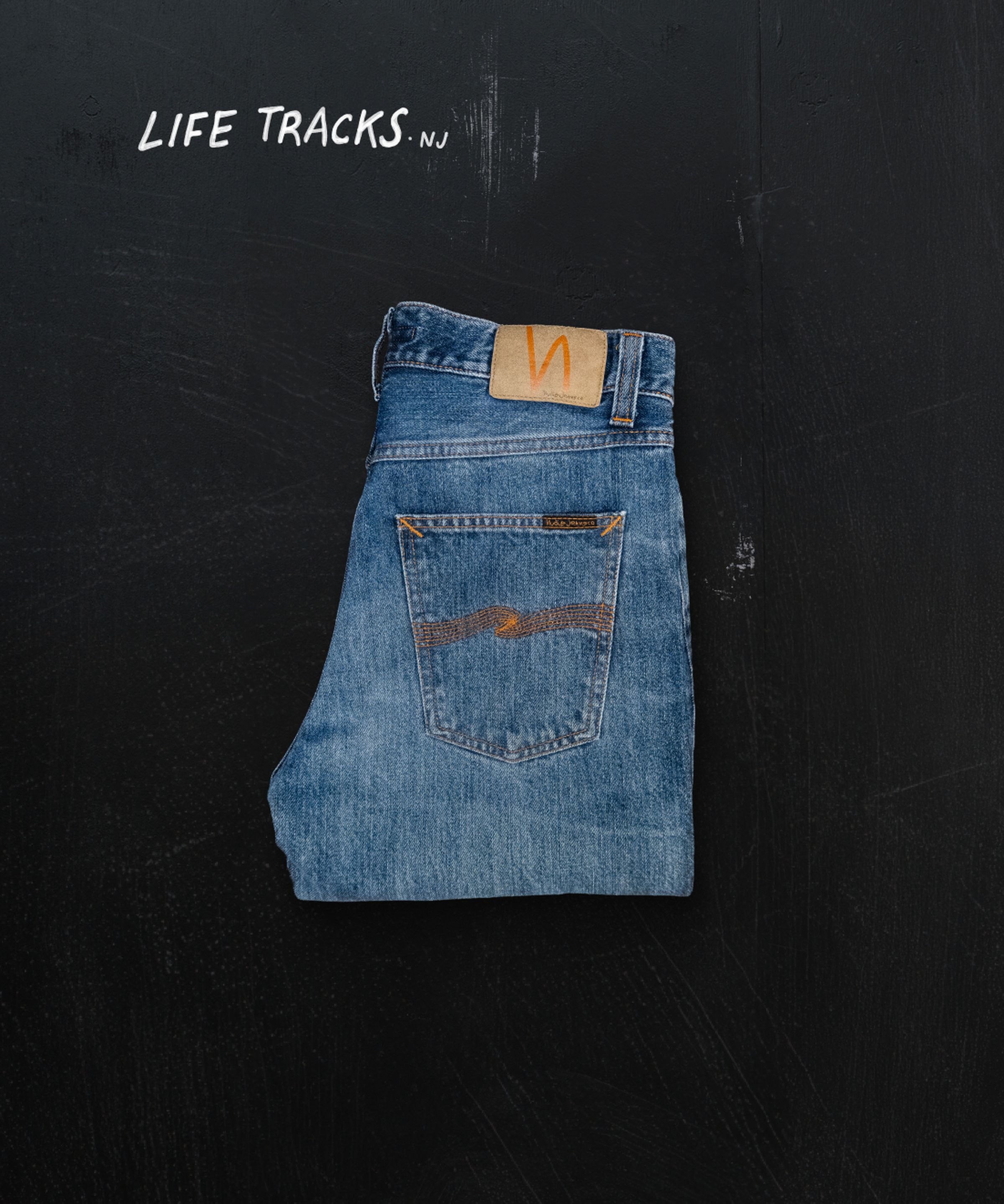 Folded blue jeans with a leather patch, placed on a dark surface. The words "LIFE TRACKS NJ" are written in the top left corner.