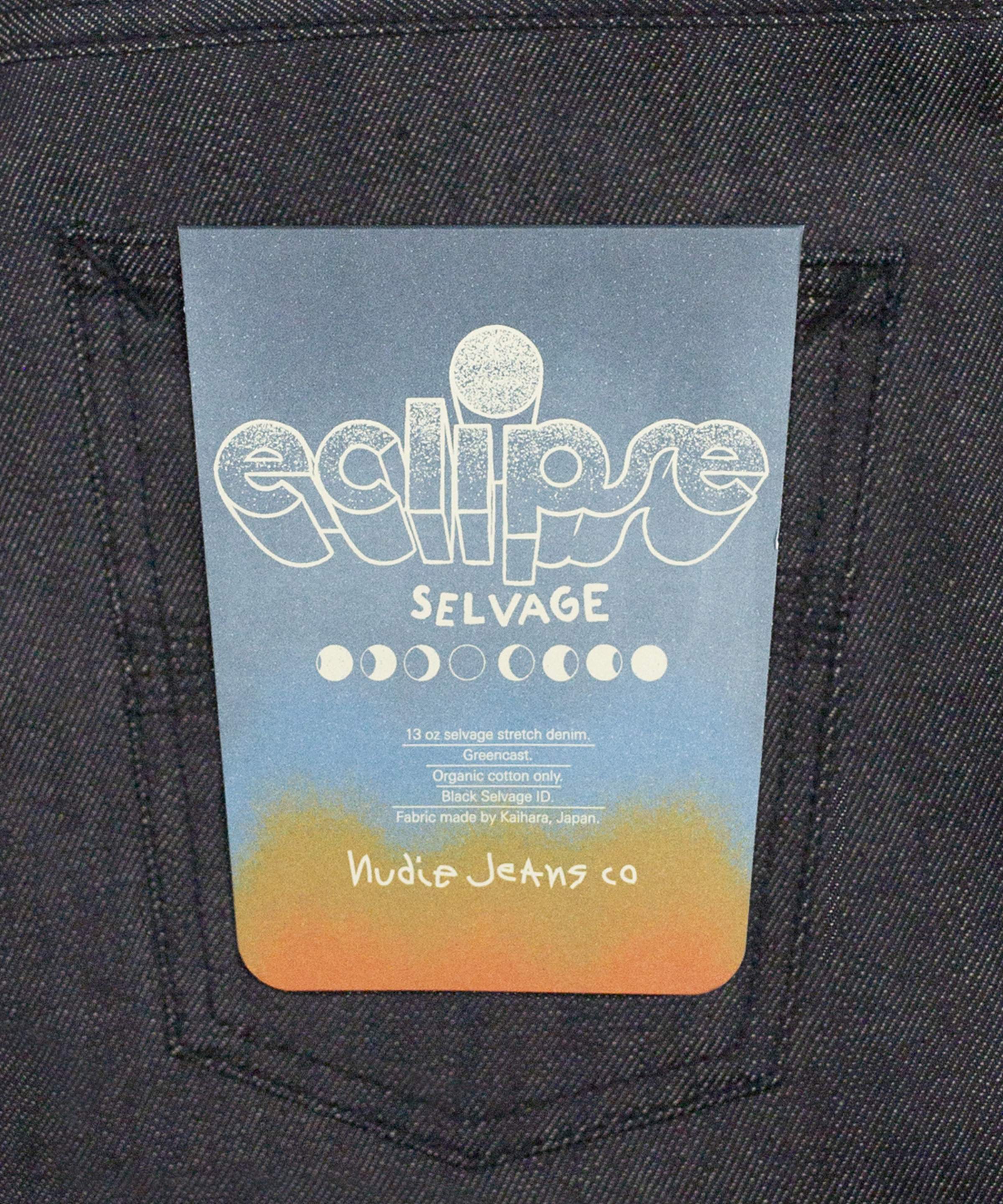 A blue and orange tag with "eclipse SELVAGE" text and moon phases design, attached to the back pocket of dark denim jeans.