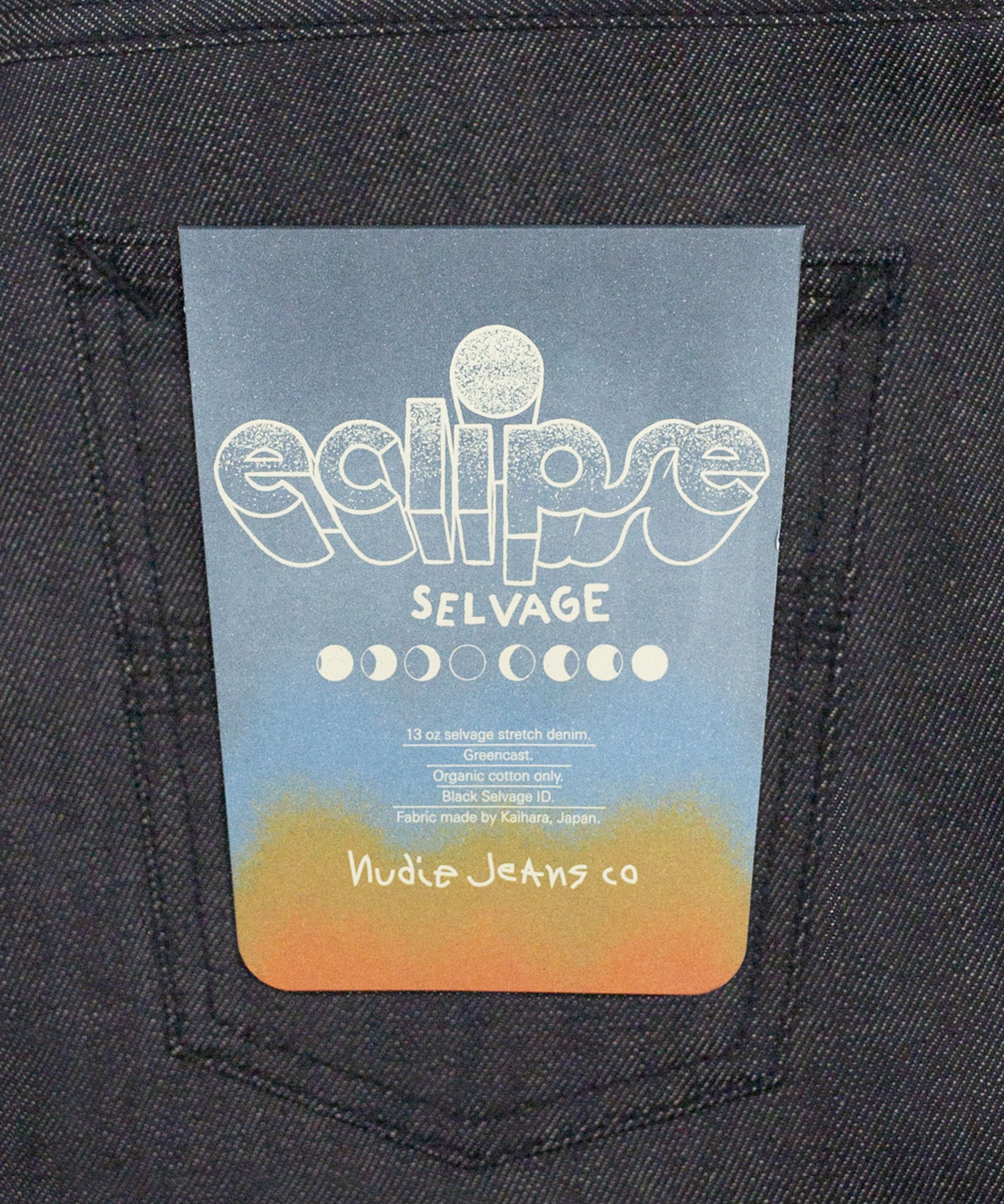 A blue and orange tag with "eclipse SELVAGE" text and moon phases design, attached to the back pocket of dark denim jeans.