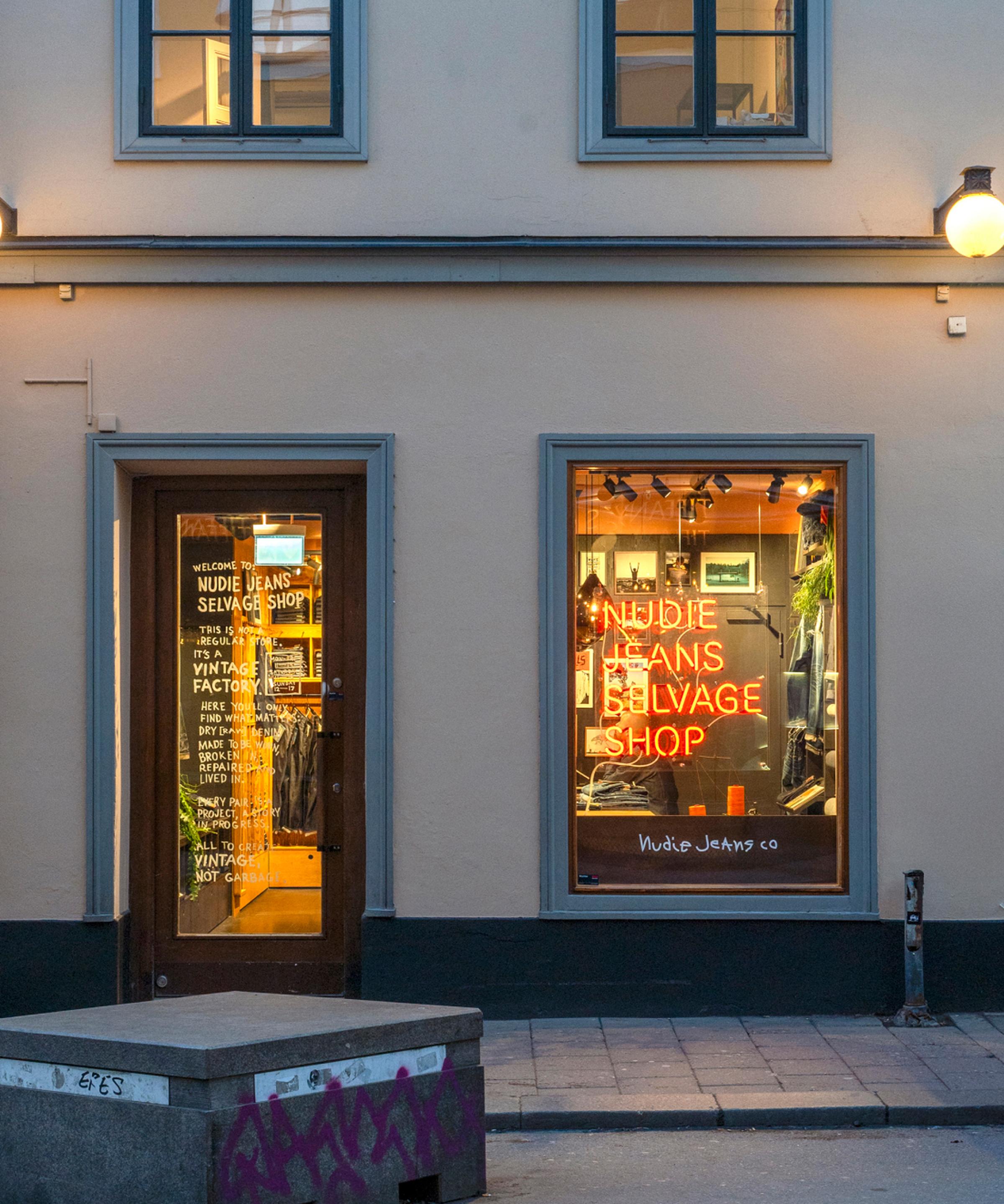 Exterior of a storefront with a wooden door and large window displaying a neon sign reading "Nudie Jeans Selvage Shop."