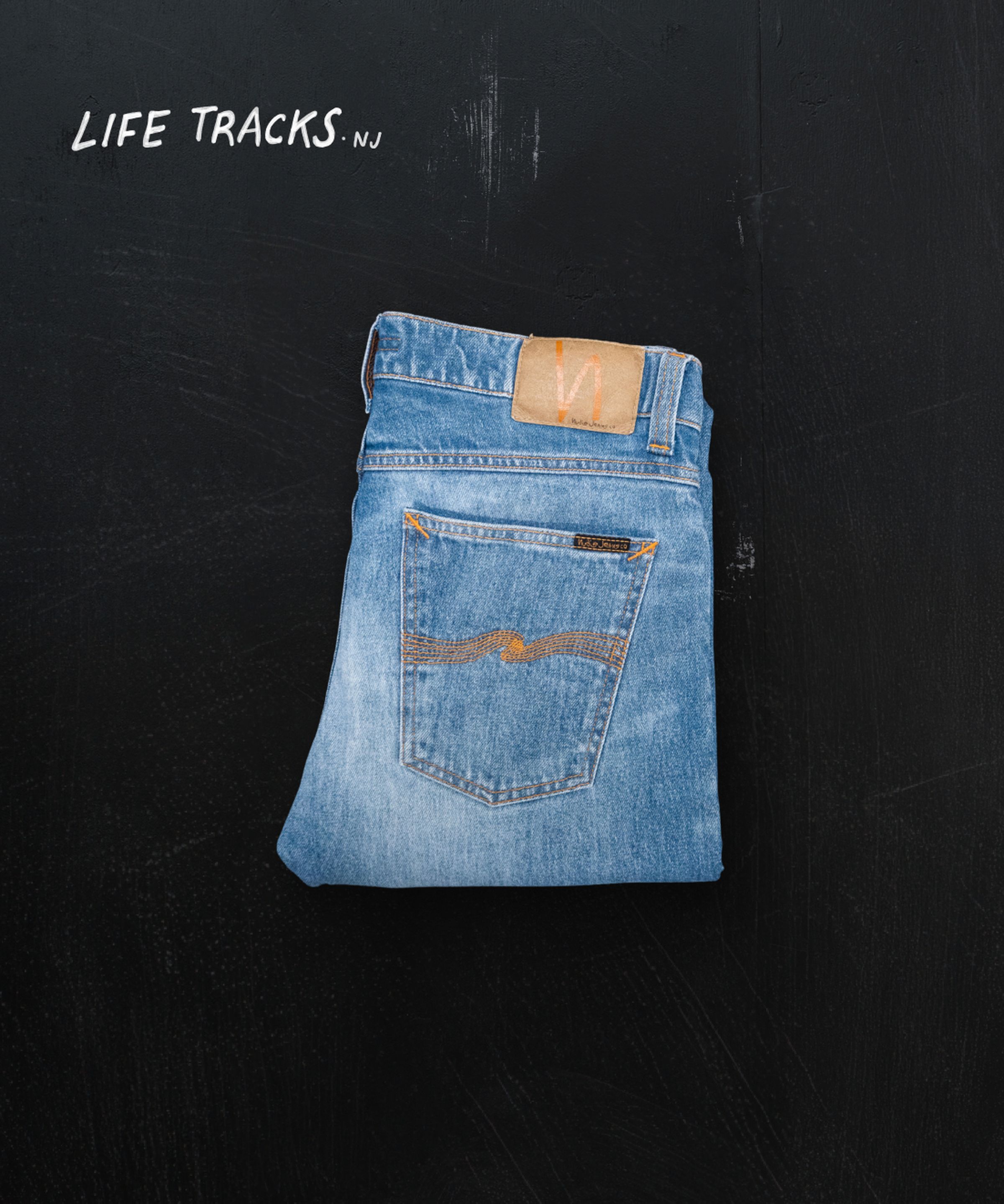 Folded blue jeans with a leather patch and orange stitching on a black background. Text reads "Life Tracks NJ" in the corner.