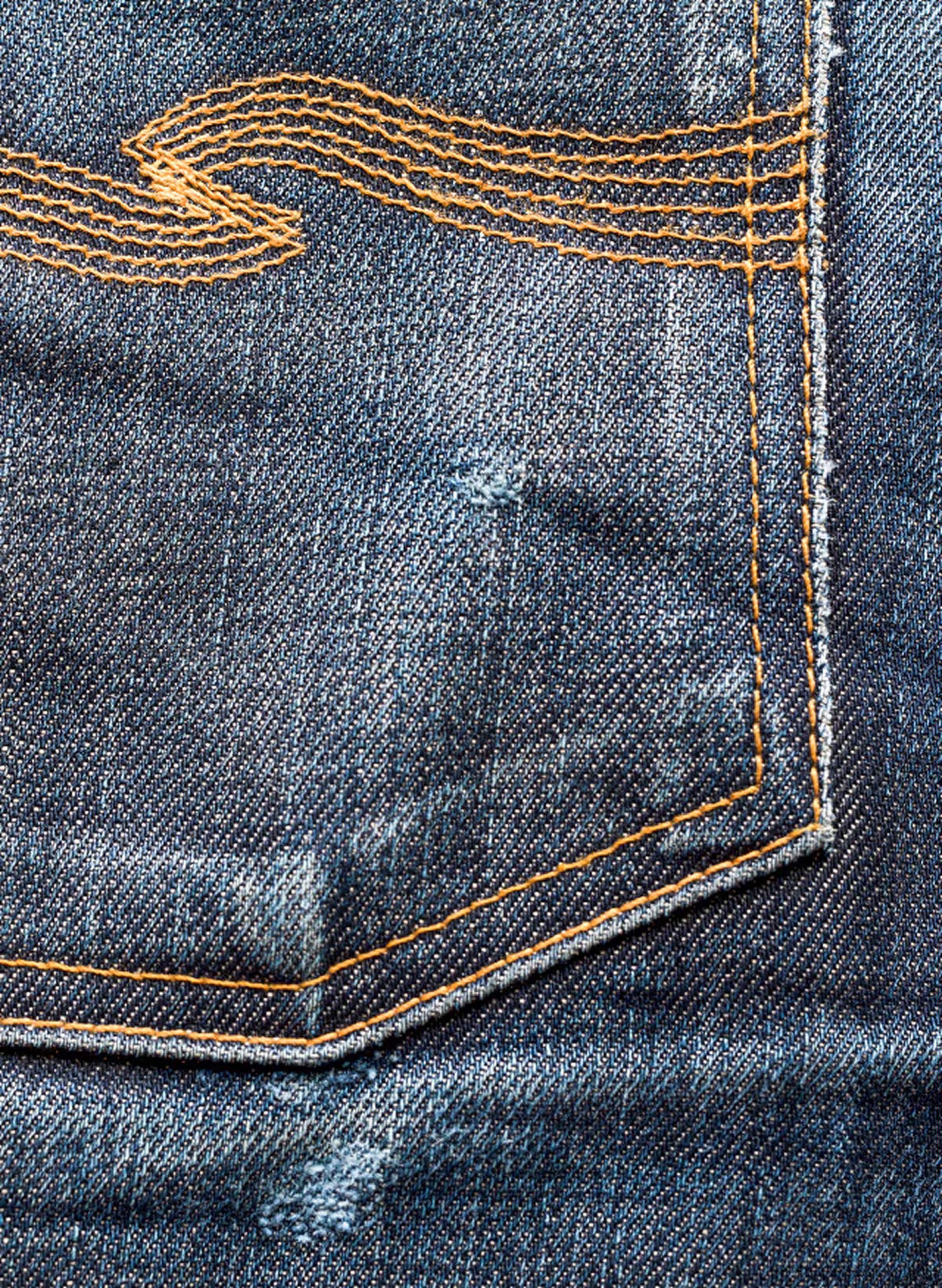 Close-up of a denim fabric with a back pocket, featuring orange stitching and a decorative wave pattern. The fabric shows slight wear and distress.