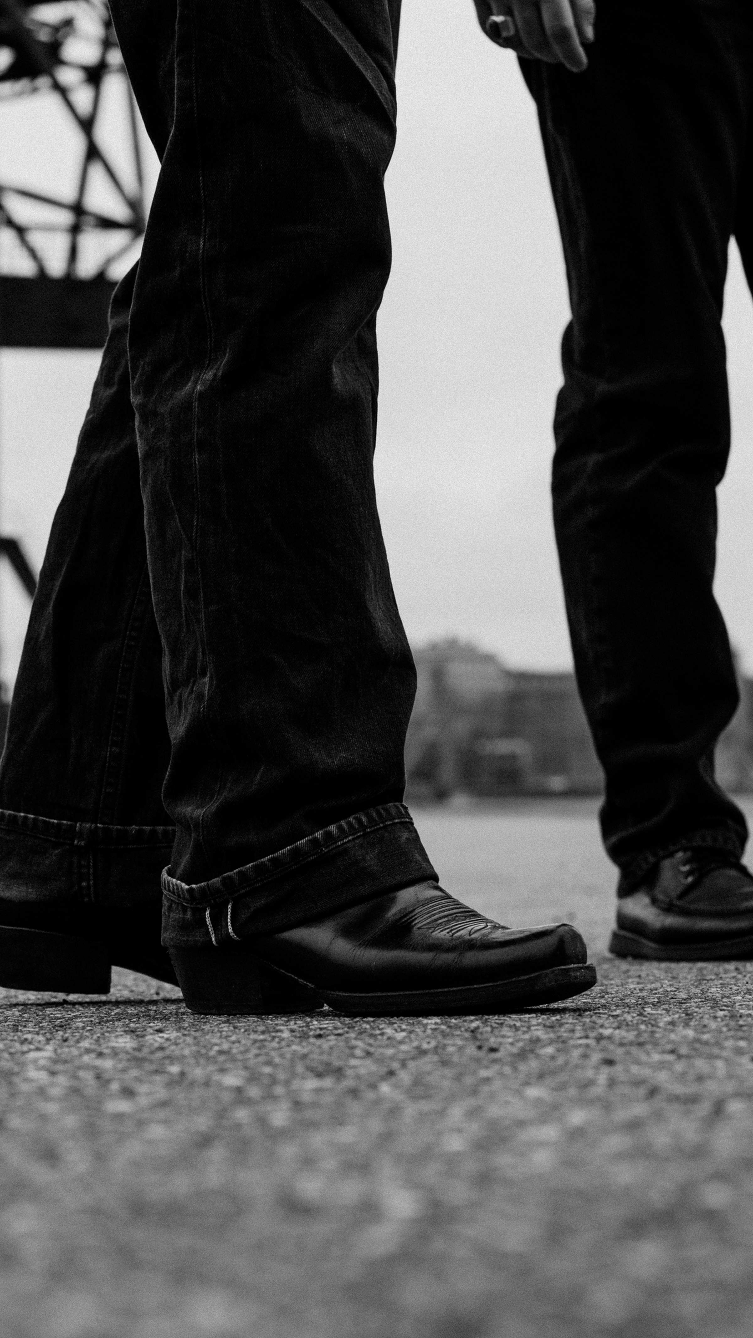 Black and white image of two people wearing dark jeans and boots, standing on a textured surface, with an industrial structure in the background.