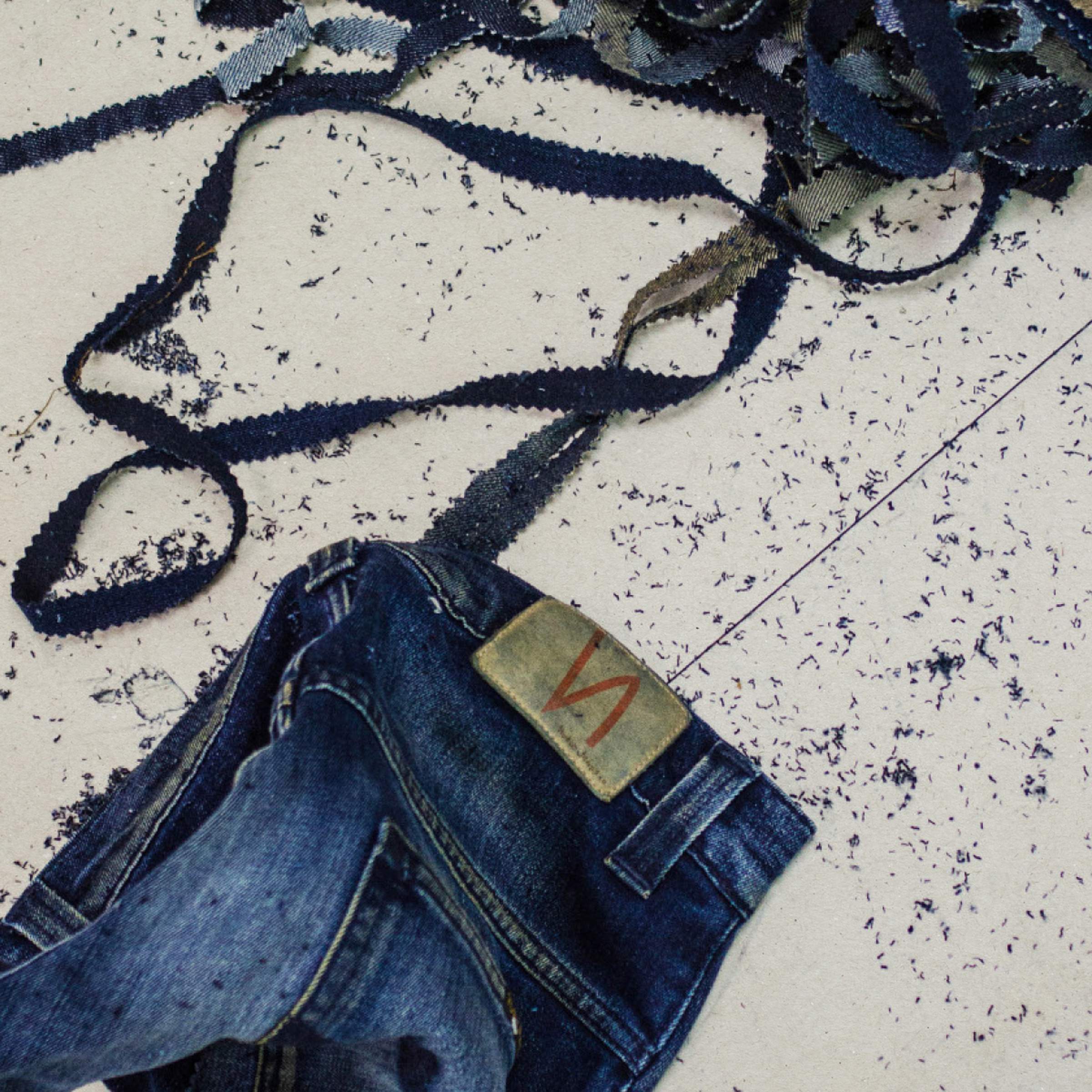 Denim strips scattered on the floor, blue jeans showing a Nudie Jeans leather patch and stitching.