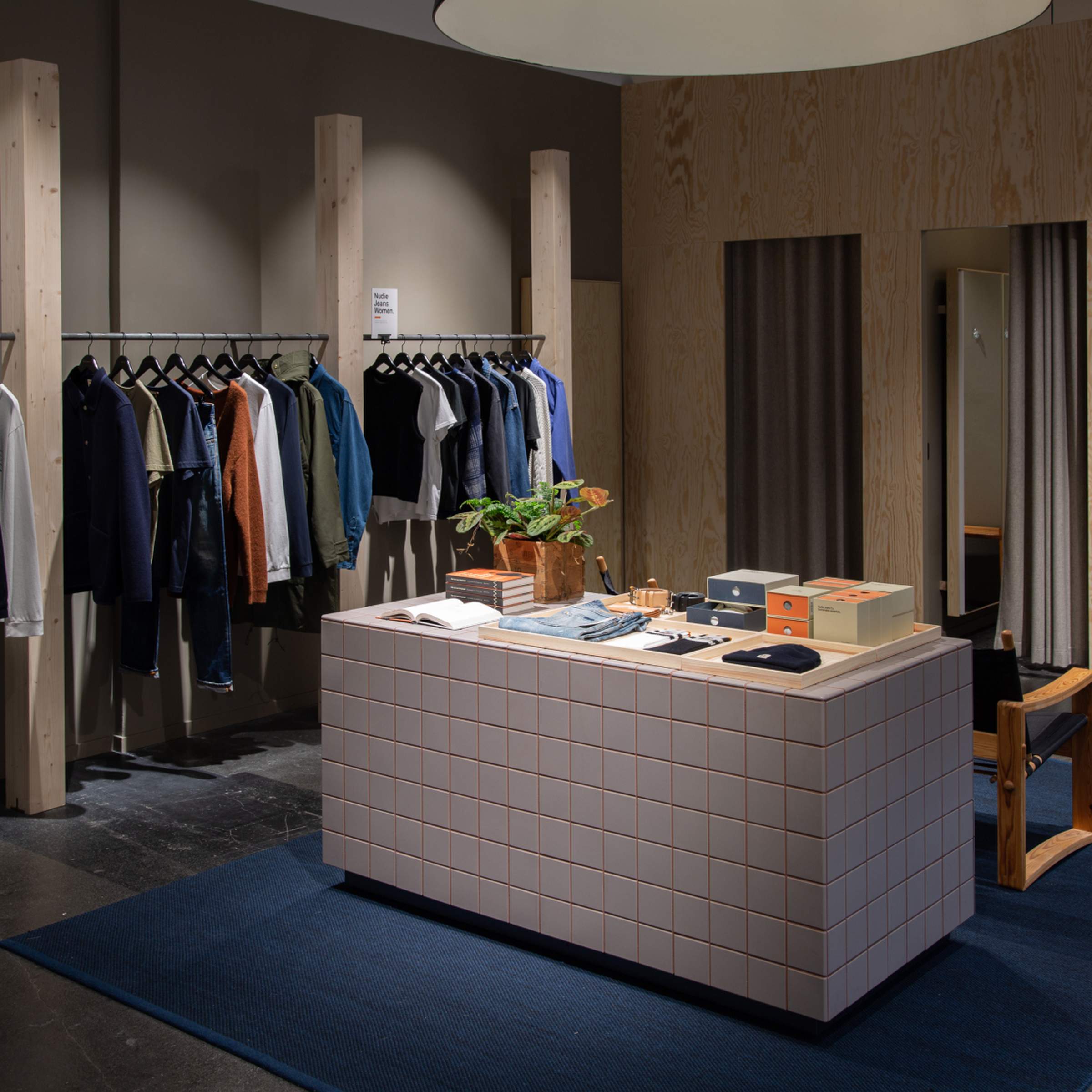 Interior of the Nudie Jeans Skånegatan Repair Shop featuring a tiled display counter, clothing racks, fitting rooms, and wooden accents.