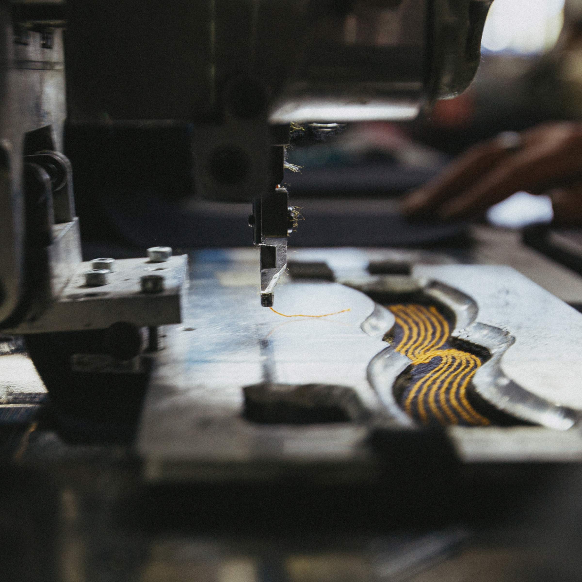 Close-up of a sewing machine needle working on denim fabric with yellow and purple thread patterns, hand in the background.