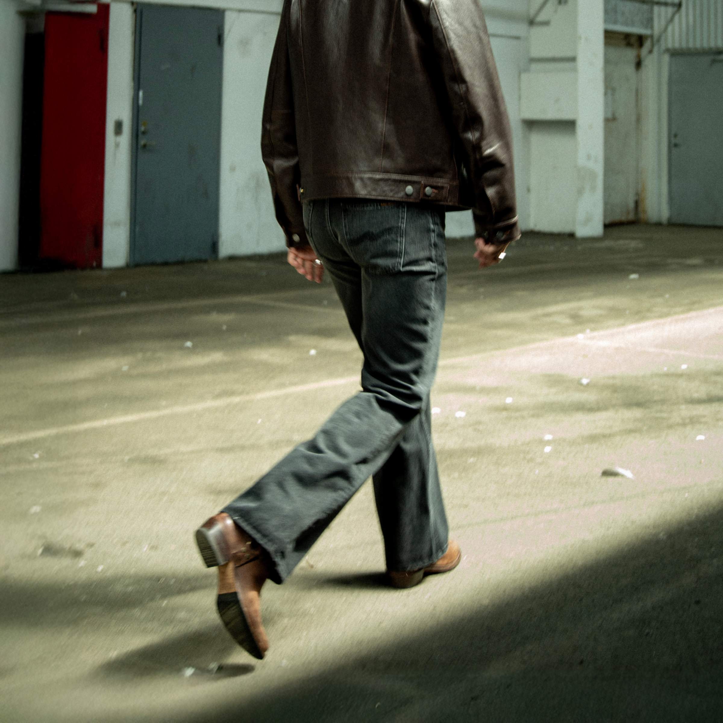 Person in a brown leather jacket and  Nudie Jeans walking away on a sunlit concrete surface, casting a shadow.
