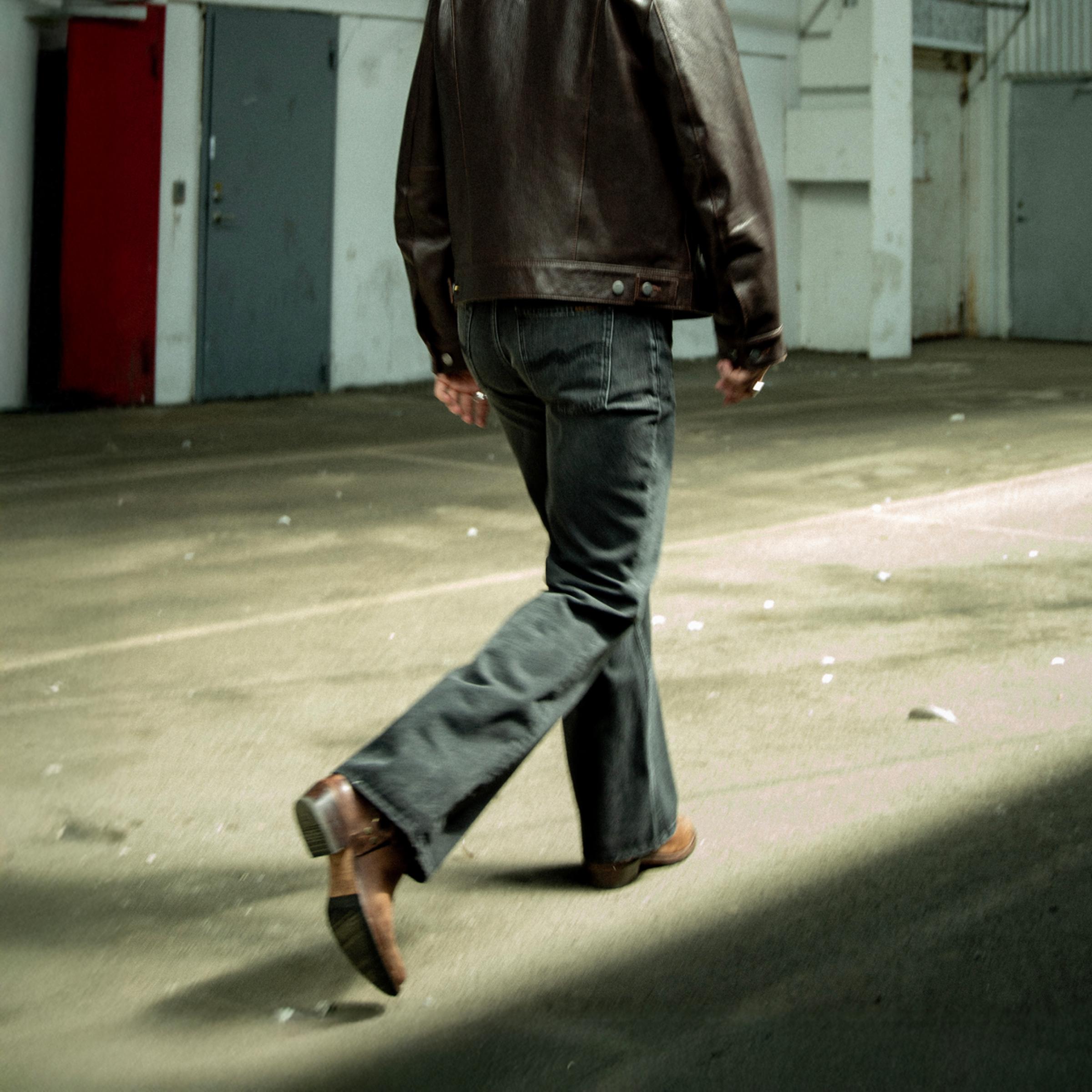 Person in a brown leather jacket and  Nudie Jeans walking away on a sunlit concrete surface, casting a shadow.