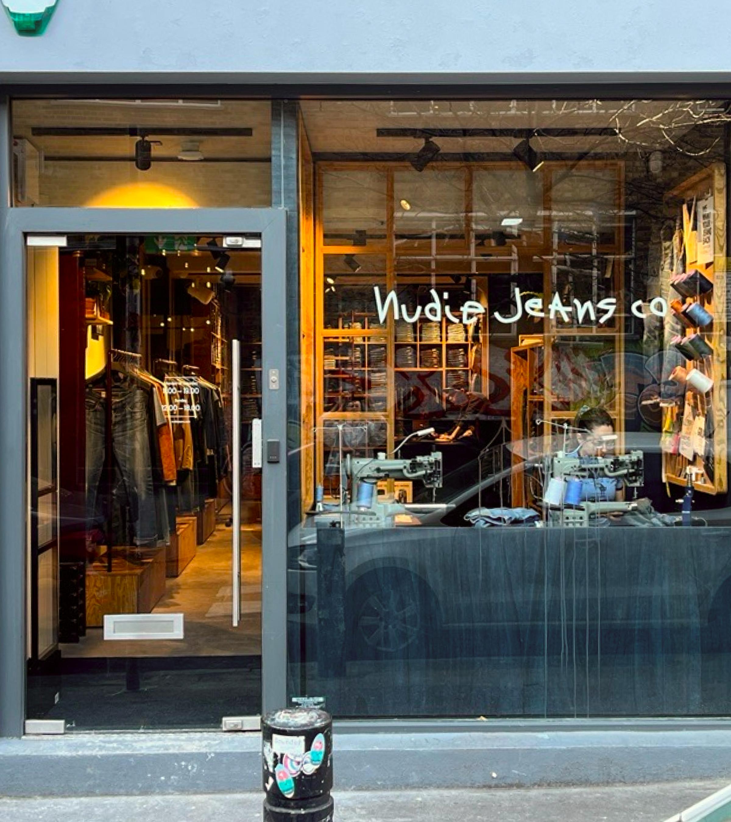 Storefront of Nudie Jeans Co. with glass door and window, showcasing denim clothing and sewing machines inside.
