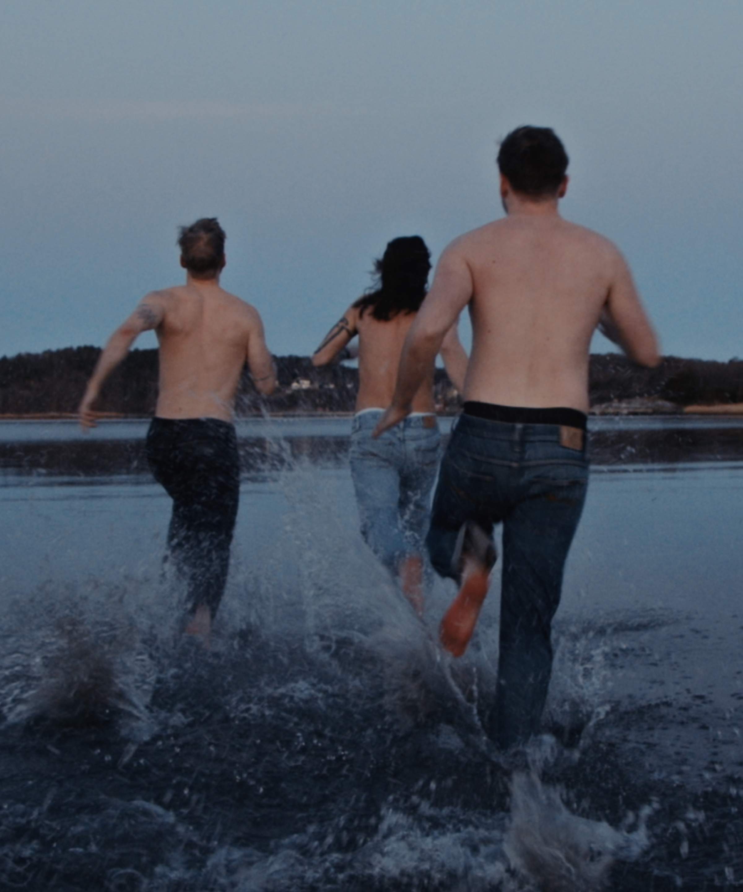 Three people in Nudie Jeans denim run shirtless into the sea at dusk, splashing water around their legs.