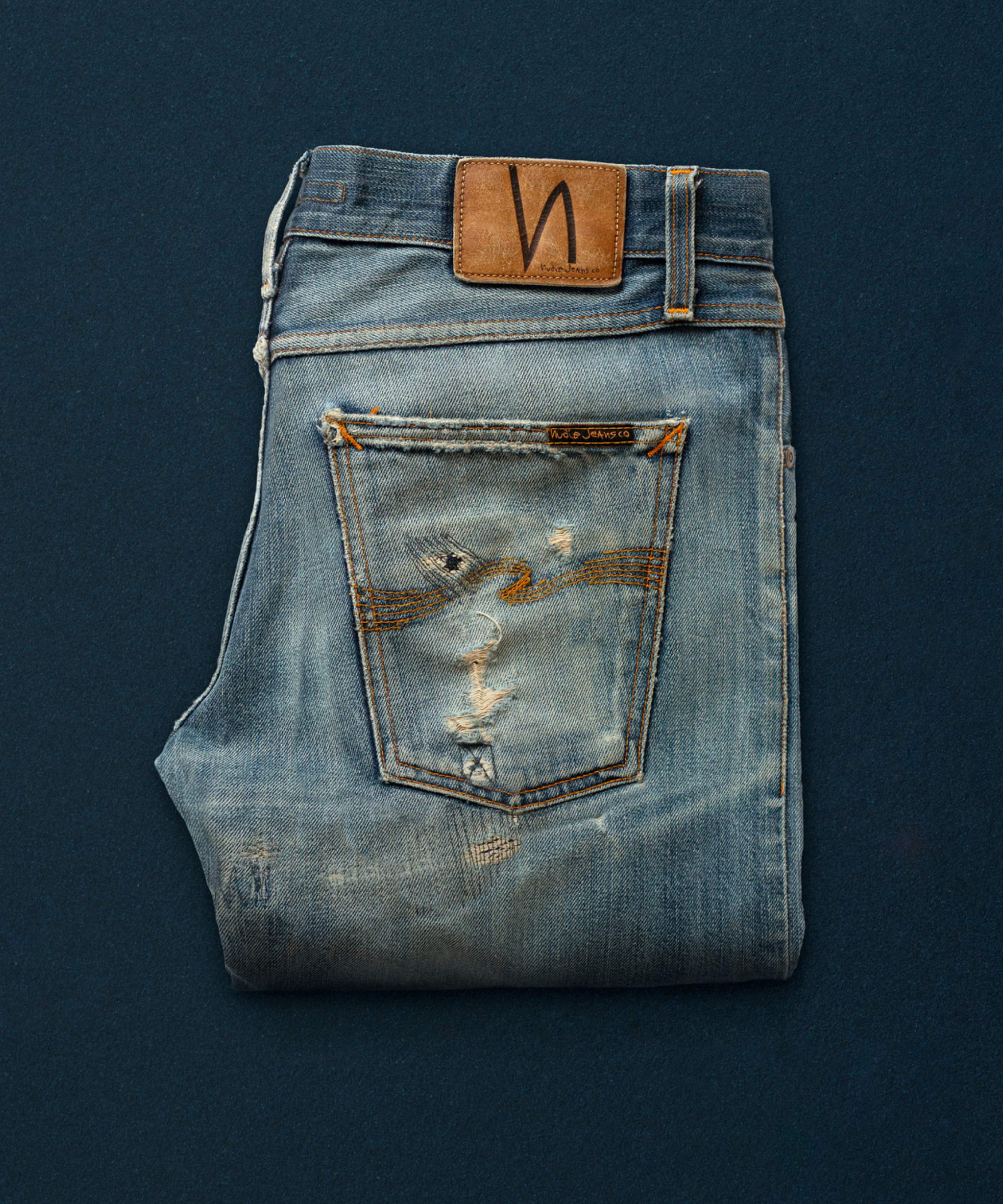A pair of well used and repaired Nudie Jeans on a blue background.