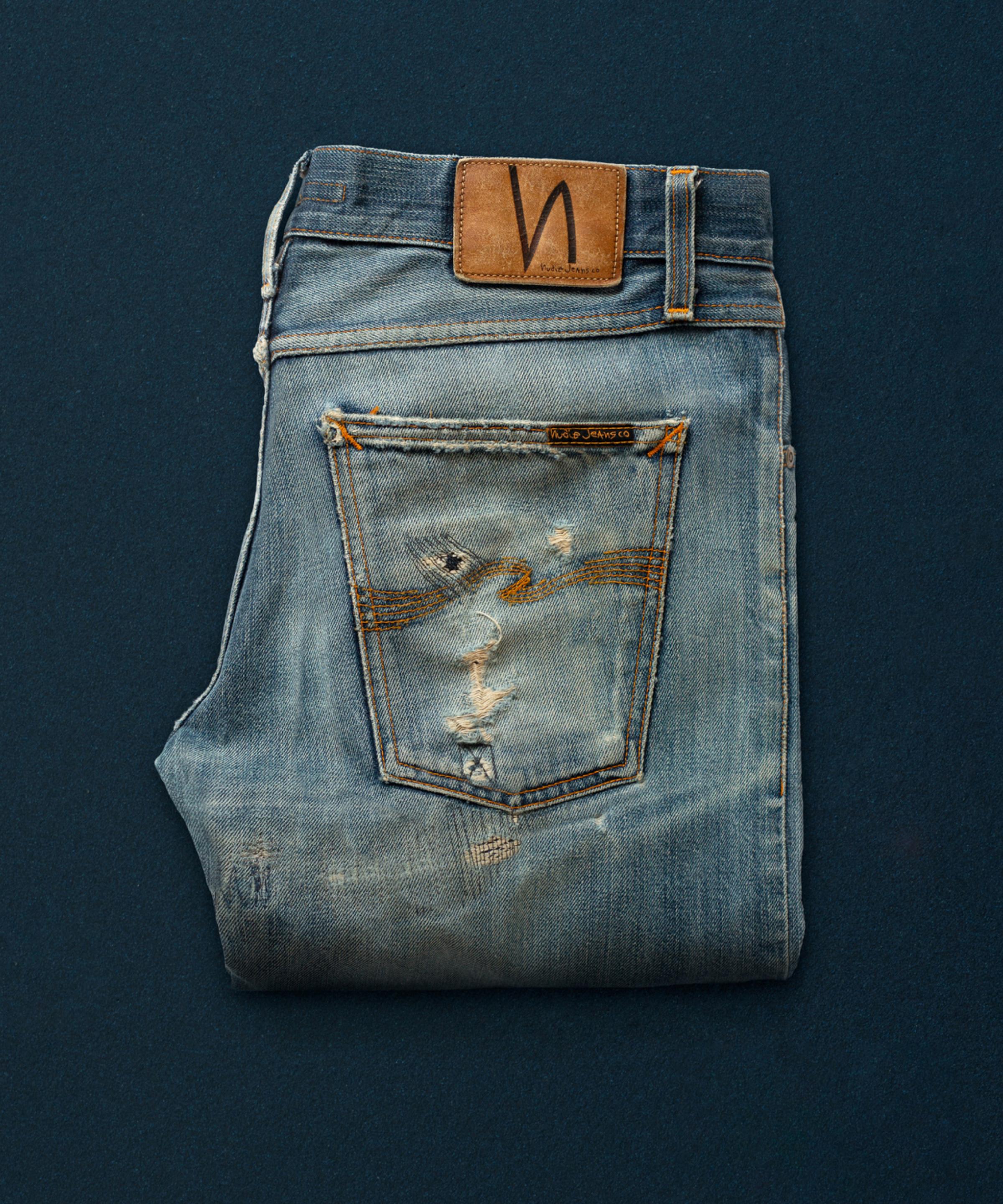 A pair of well used and repaired Nudie Jeans on a blue background.