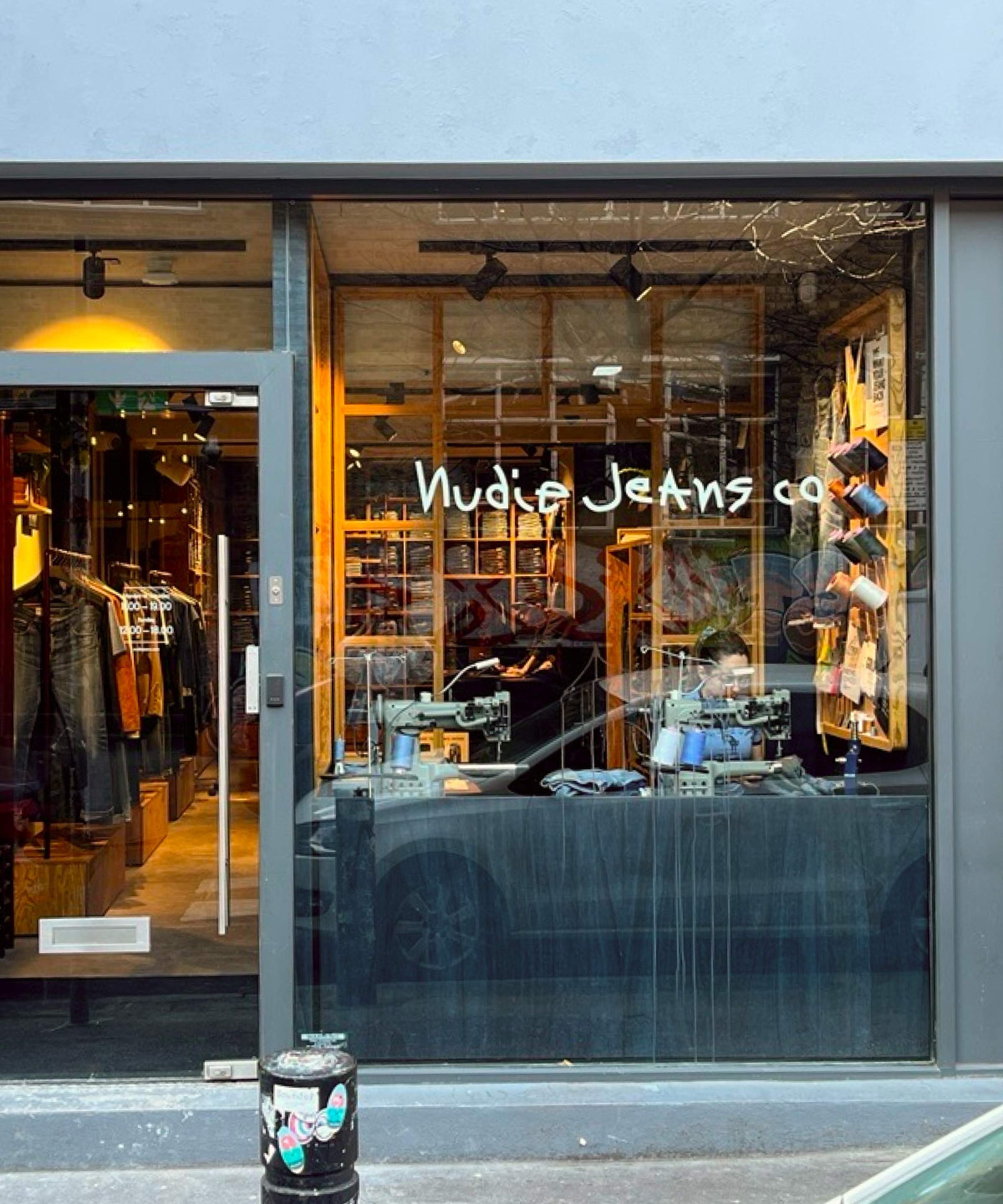 Storefront of Nudie Jeans Co with glass windows displaying clothes inside and a person working at a desk.