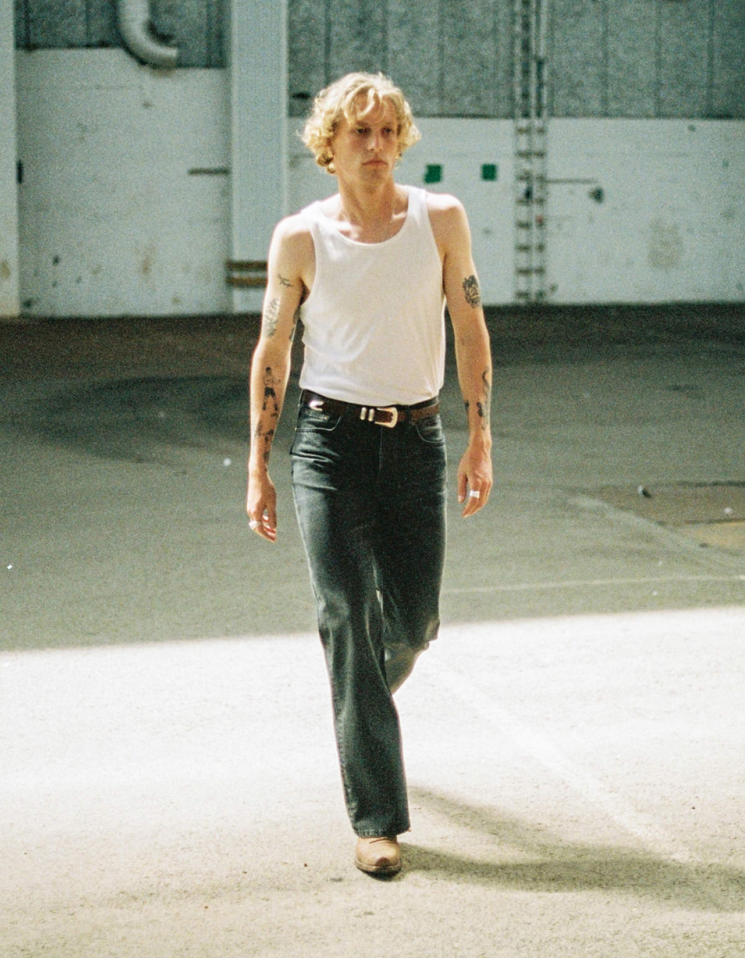 Person with curly hair, wearing a white tank top and jeans, walking confidently in a sunlit industrial space. Tattoos visible on arms.