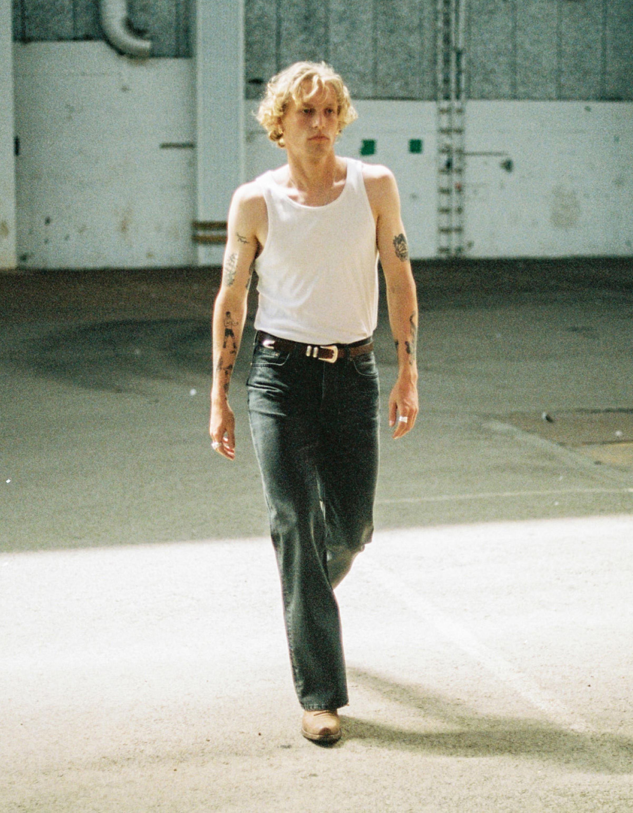 Person with curly hair, wearing a white tank top and jeans, walking confidently in a sunlit industrial space. Tattoos visible on arms.