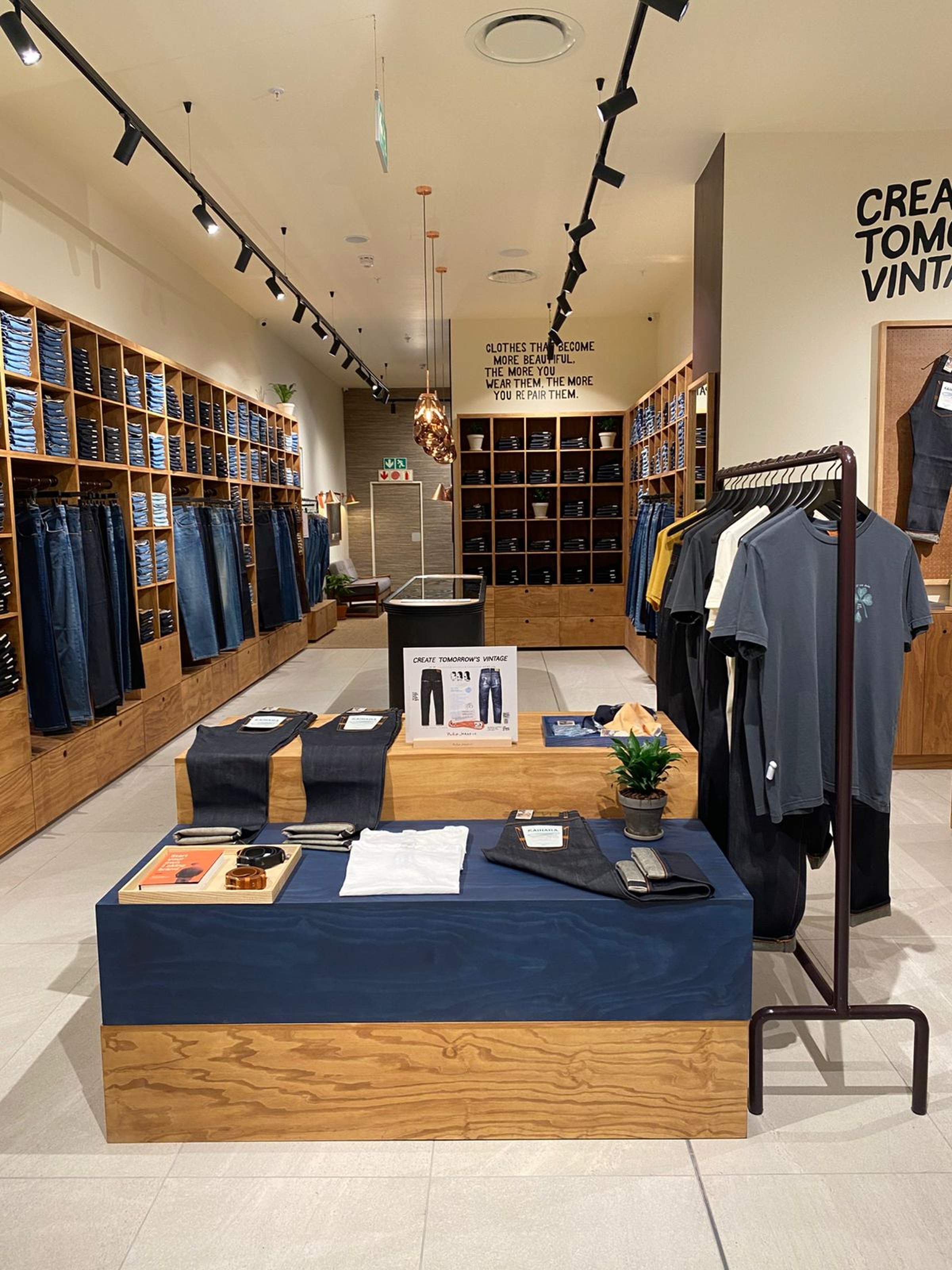 Clothing store interior with wooden shelves displaying jeans and shirts, tables with folded clothes, and a rack with hanging T-shirts.