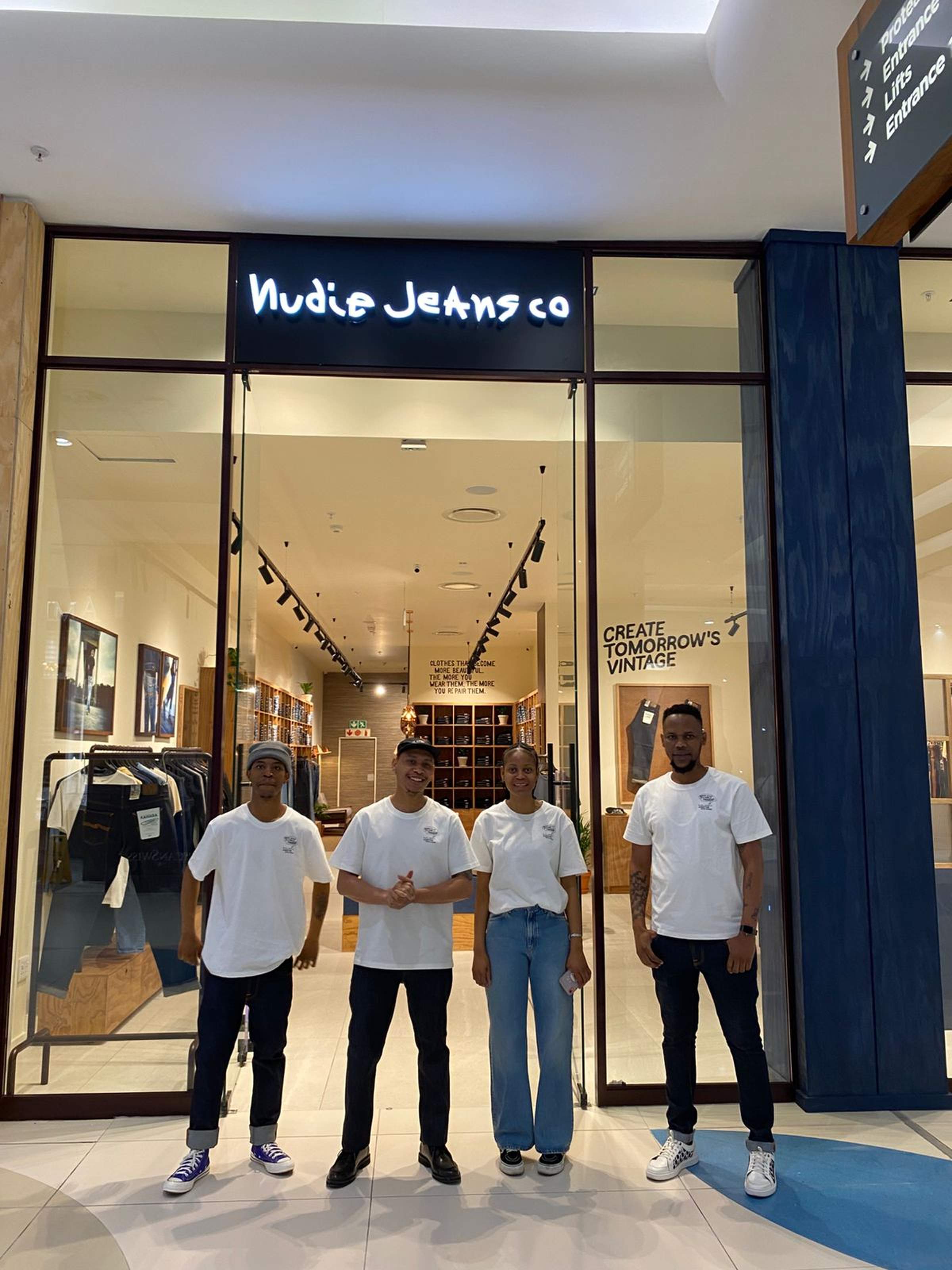 Four people in white shirts stand smiling in front of a Nudie Jeans Co store with glass doors and a sign reading "Create Tomorrow's Vintage."