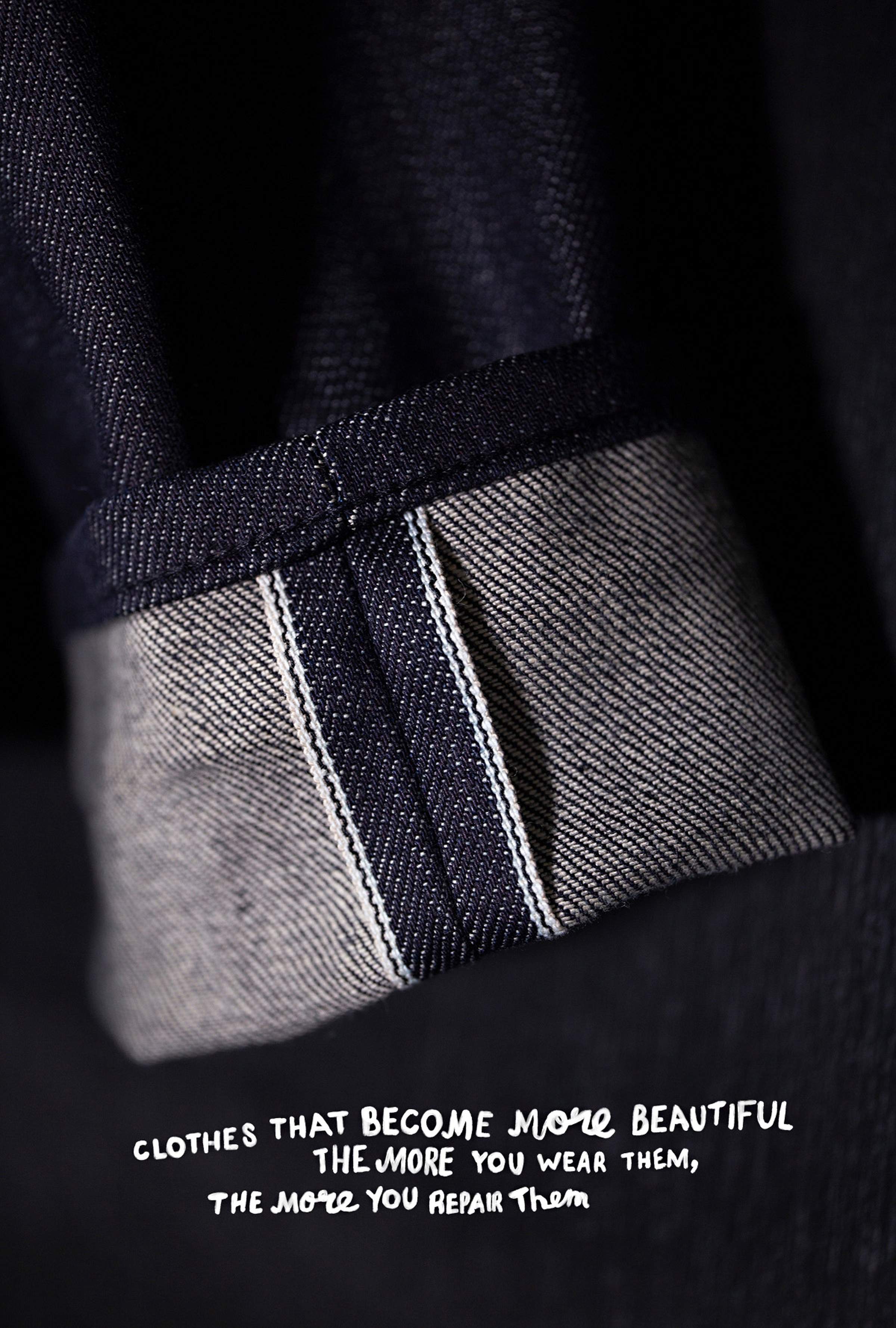 Close-up of a dark denim fabric with a folded cuff, showing detailed stitching. Text reads: "Clothes that become more beautiful the more you wear them, the more you repair them."