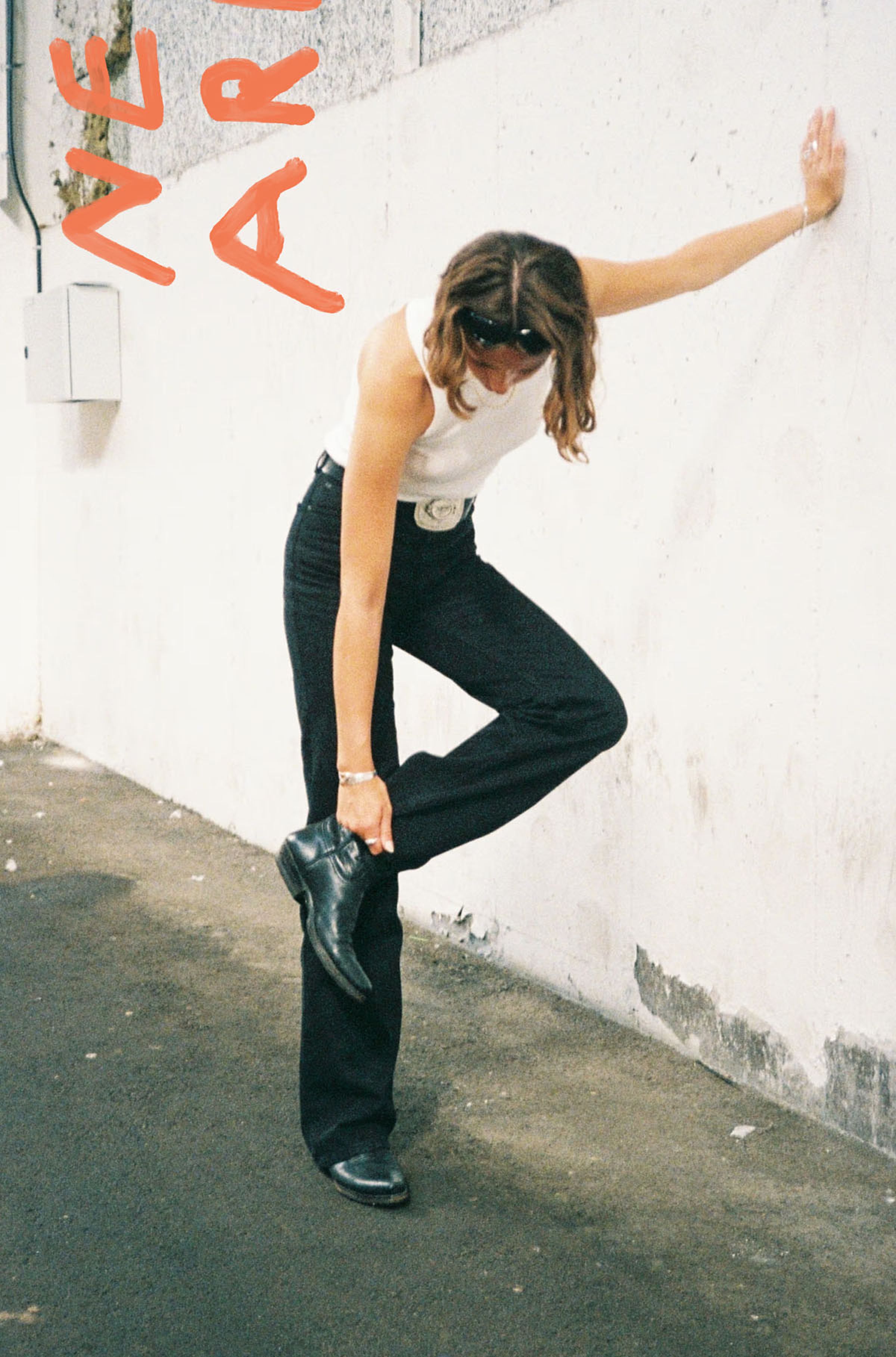 Woman in a white tank top and black pants leans against a wall, adjusting her black boot. Red text on the wall reads "NEAR."