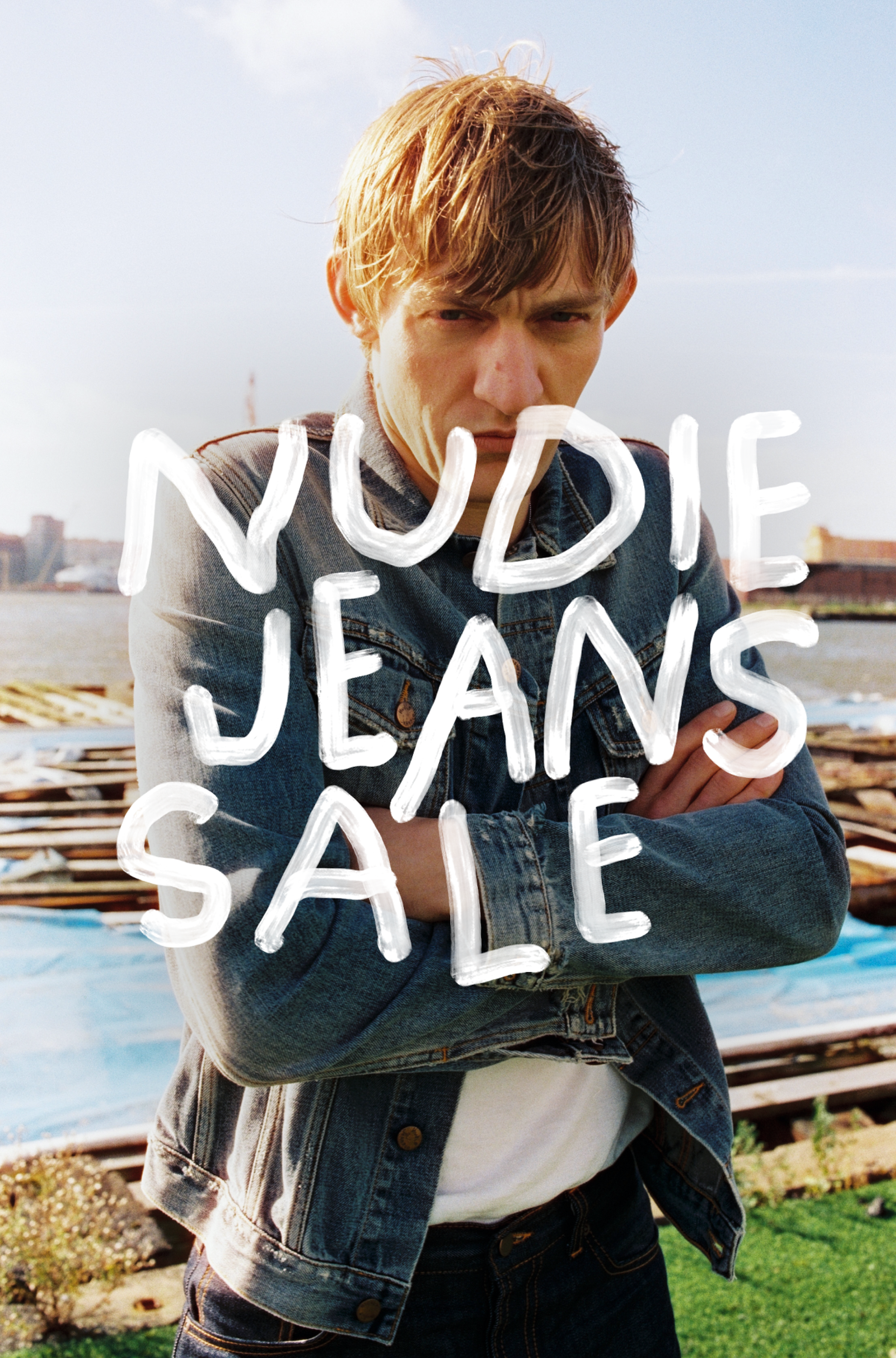 Person standing outdoors in a denim jacket with arms crossed, overlaid with the text "Nudie Jeans Sale." Water and cityscape in the background.