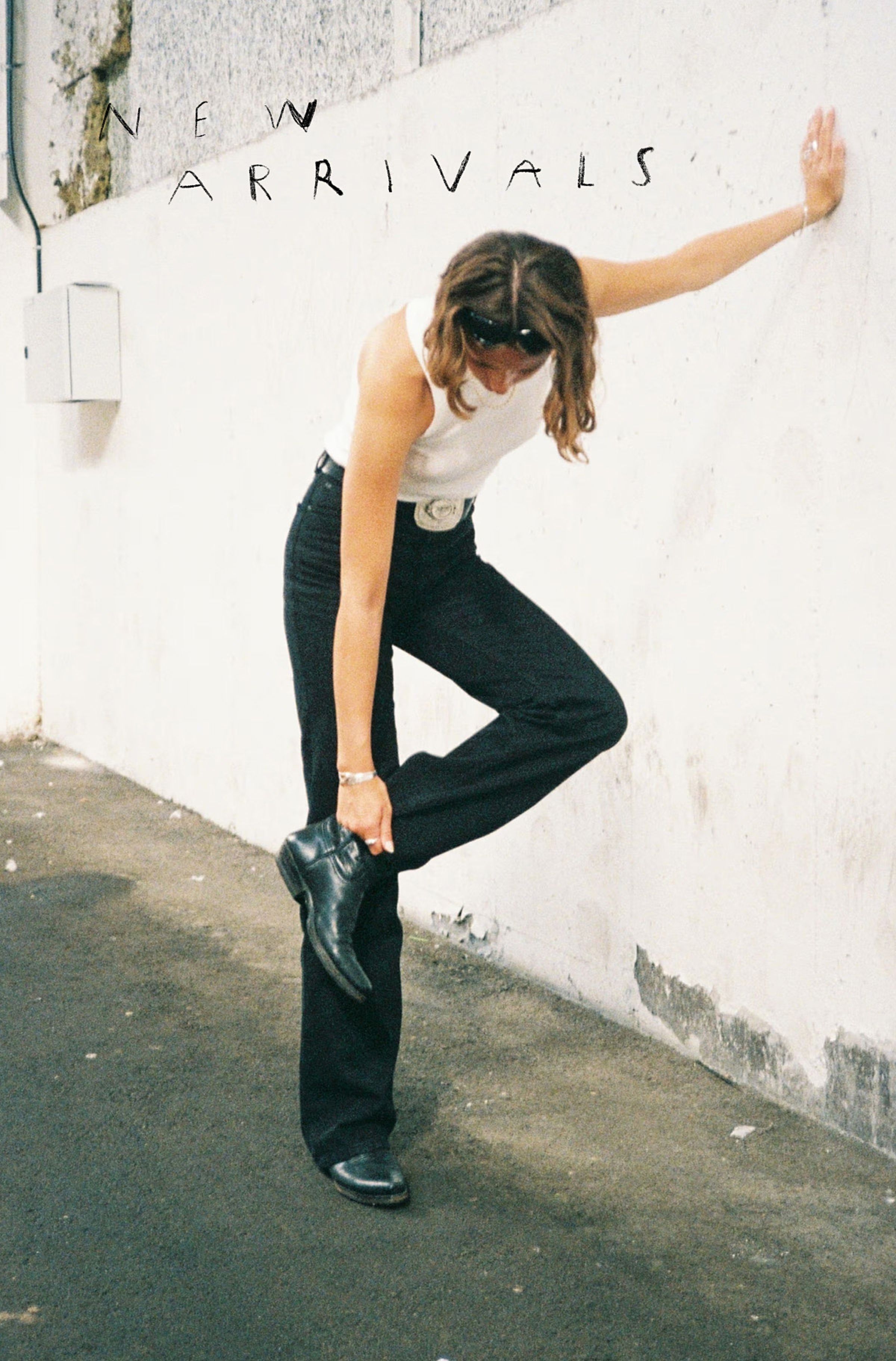 Woman in a white tank top and black pants adjusts her boot, leaning against a wall with "New Arrivals" text above.