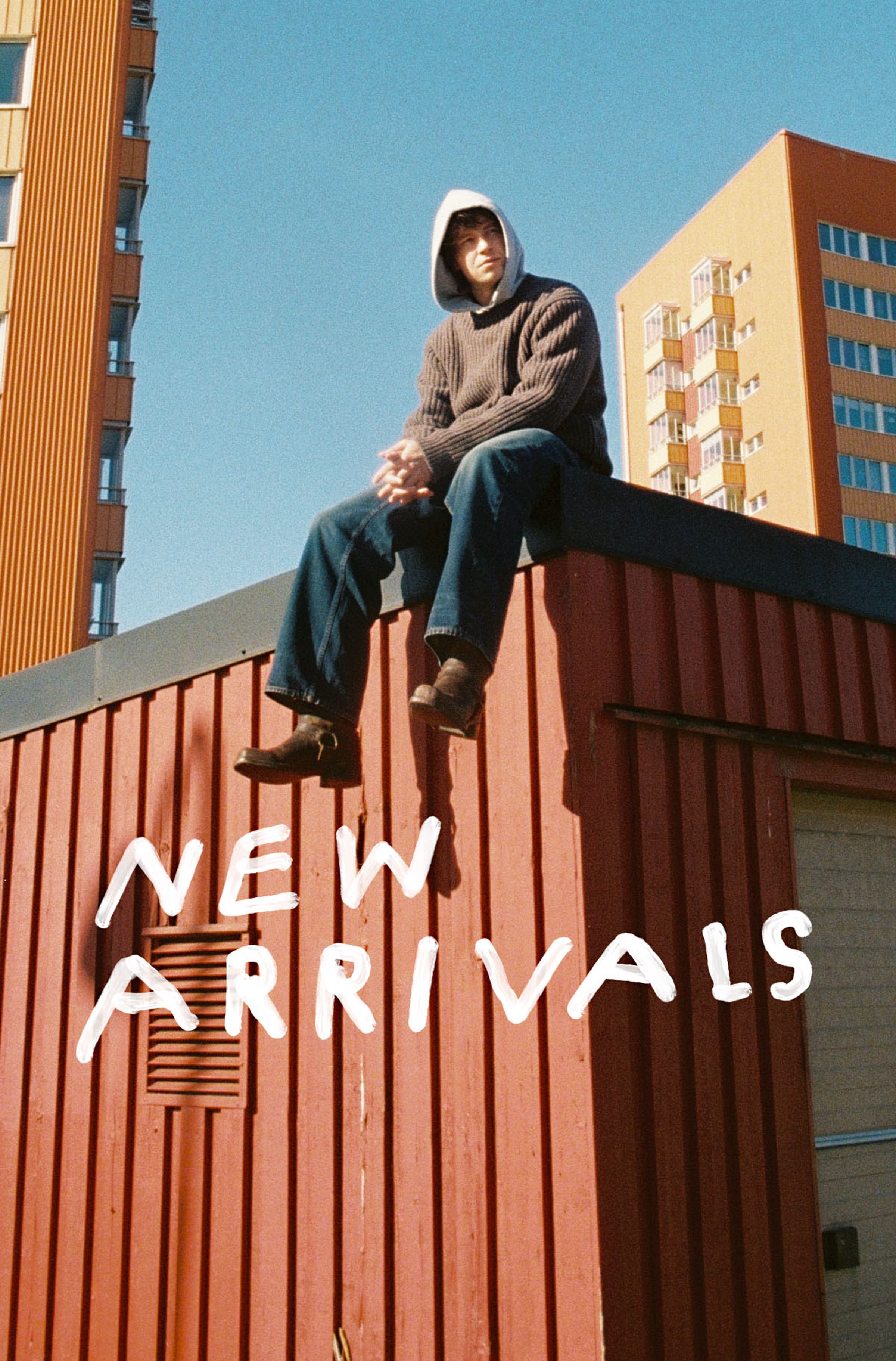 Person in a hoodie and and Nuide Jeans sits on a red rooftop with modern buildings in the background. Text reads "New Arrivals."