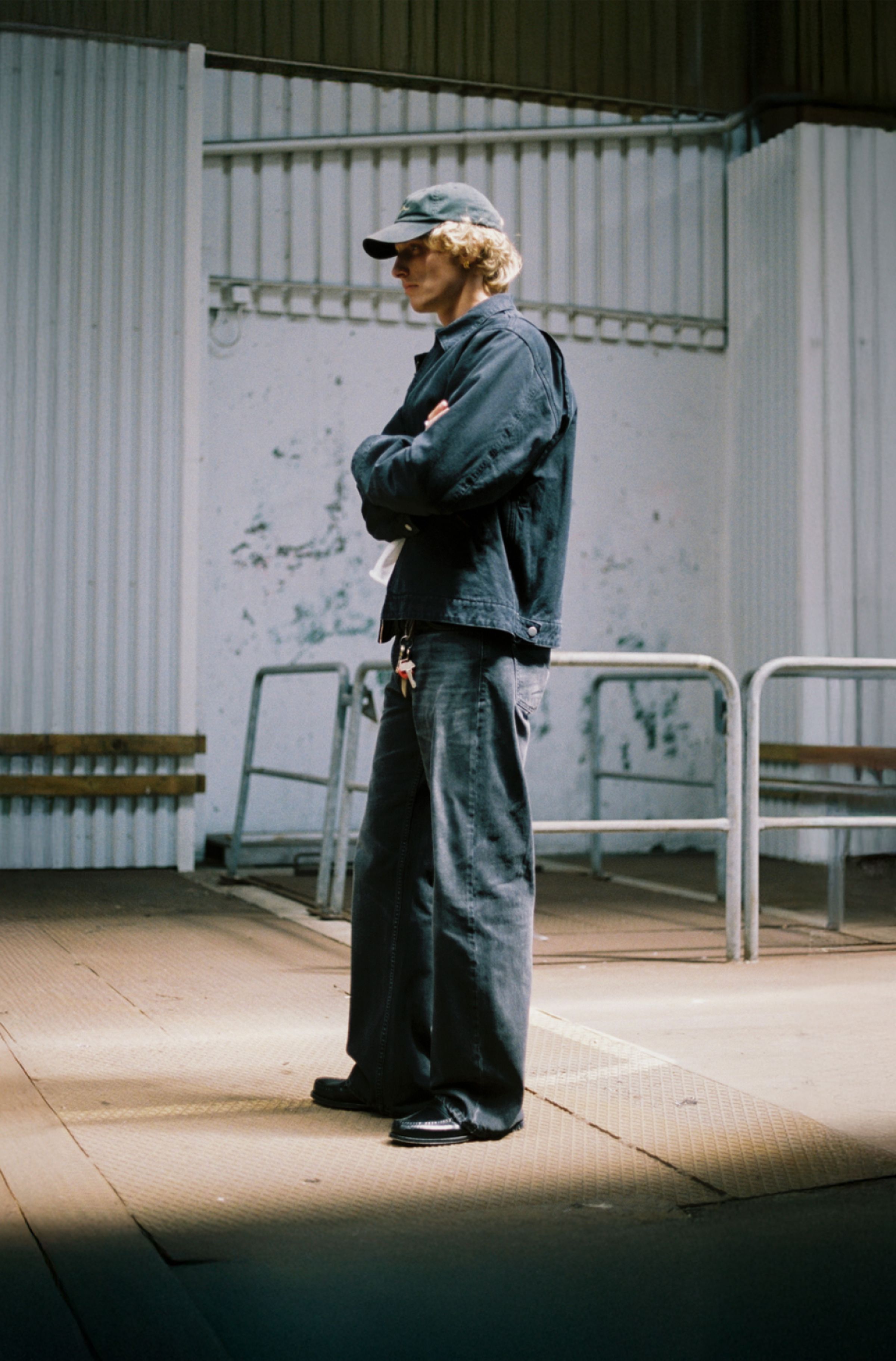 Person in a black jacket and cap standing with arms crossed in an industrial setting with metal railings and white walls.