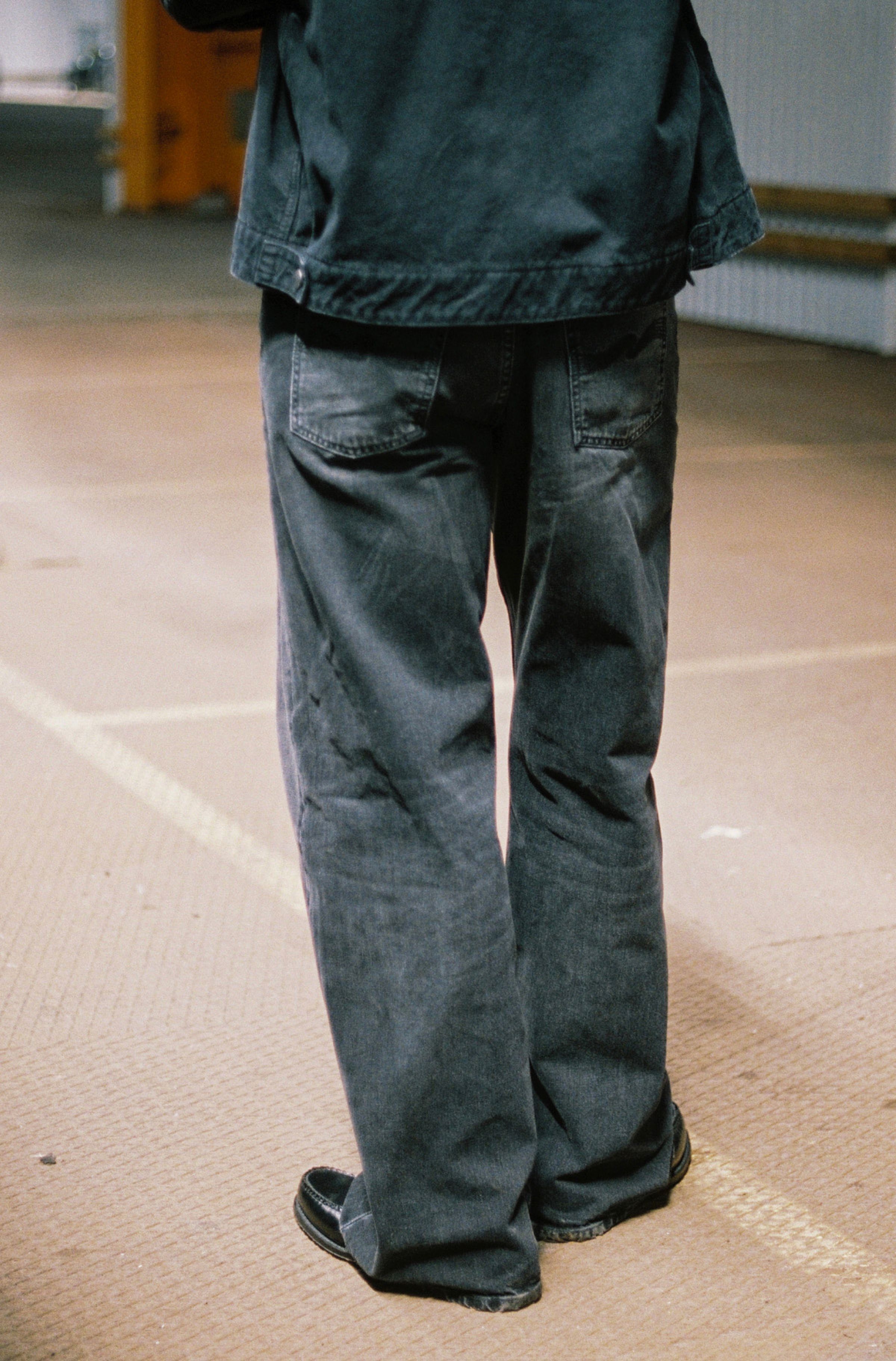 Person wearing black jeans and a dark jacket, standing on a beige floor with a blurred background.